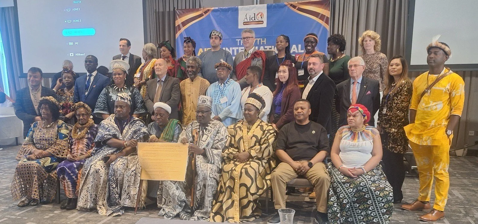 Group photo of diverse individuals in traditional and modern attire at an international conference, with a blue banner and projection screens in the background.