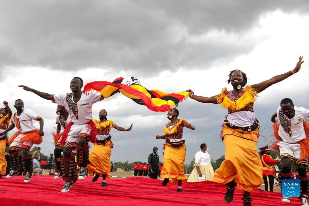 People dressed in colorful traditional African clothing dancing on a red stage outdoors under a cloudy sky, with two women holding a flag.