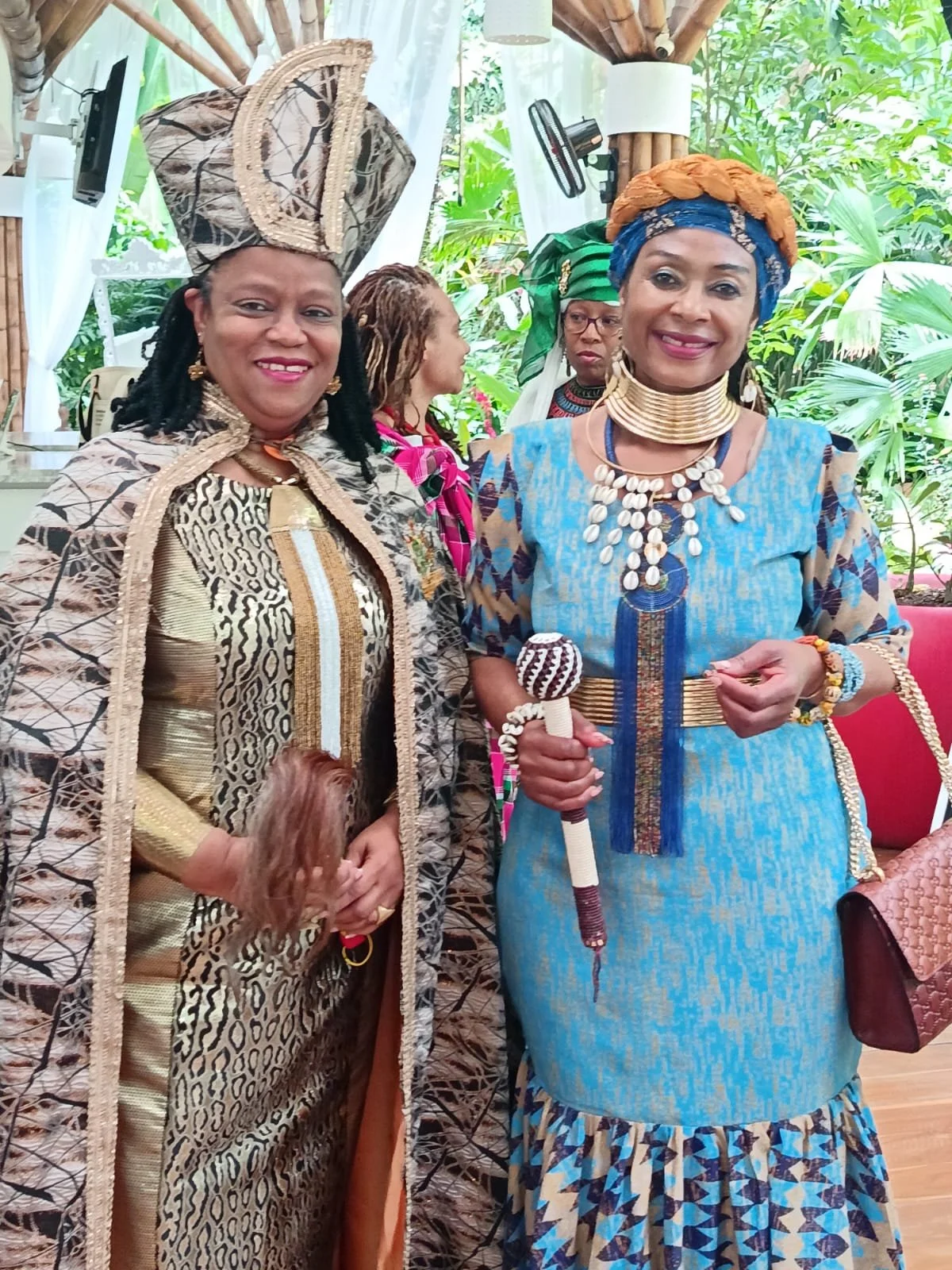 Several women dressed in traditional African attire, wearing jewelry and headpieces, at a cultural gathering. Two women in the foreground are smiling, with one holding beaded accessories and a staff.
