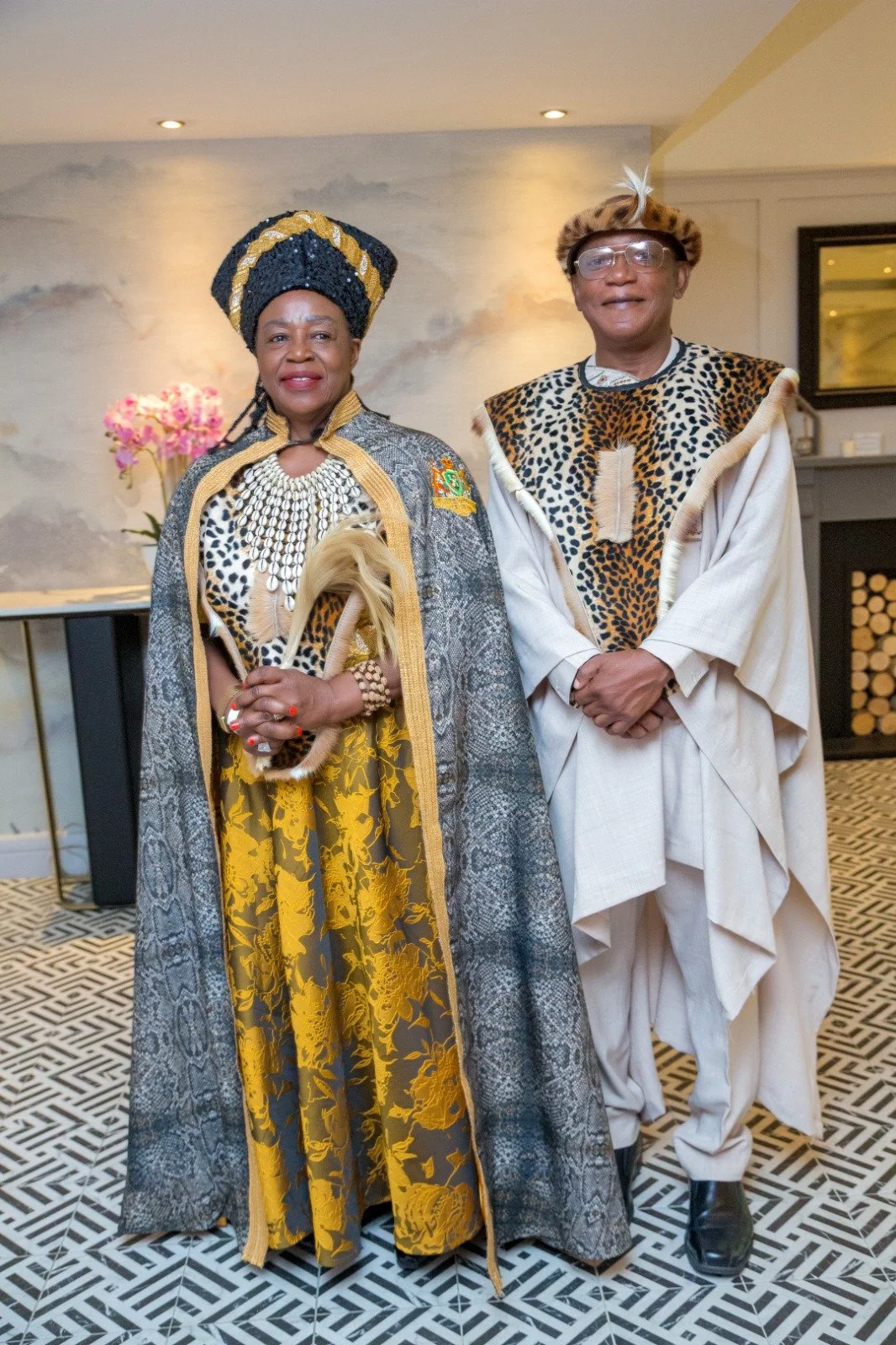 A woman and a man dressed in traditional African attire, standing indoors with patterned flooring and decorated with flowers in the background.