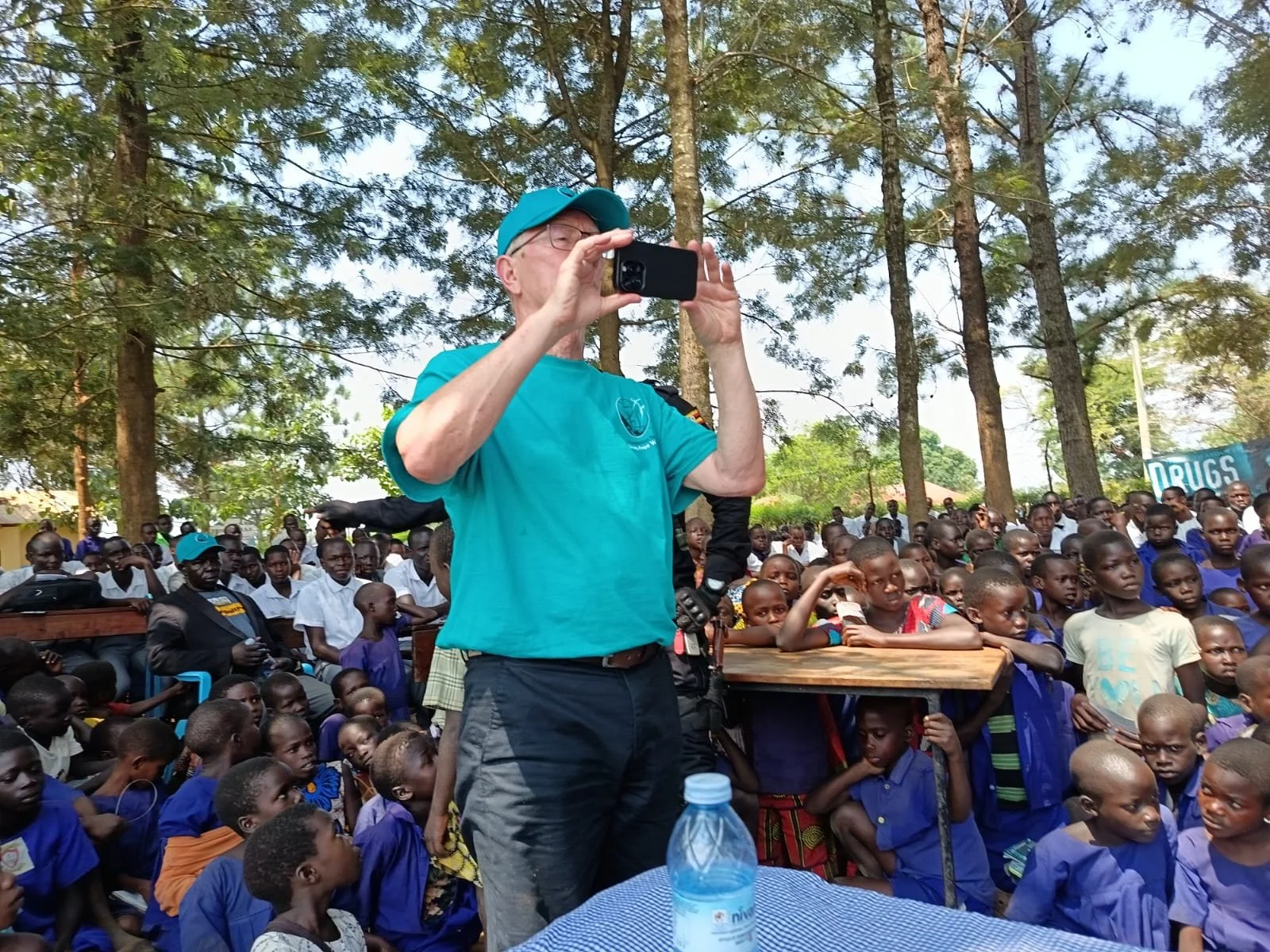 A man in a blue shirt and cap taking a photo or recording video with a smartphone in front of a crowd of children and adults sitting outdoors under trees at a community event.