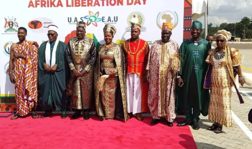 Group of nine people dressed in traditional African attire standing on a red carpet in front of a banner with the words "Africa Liberation Day" and logos of various organizations.