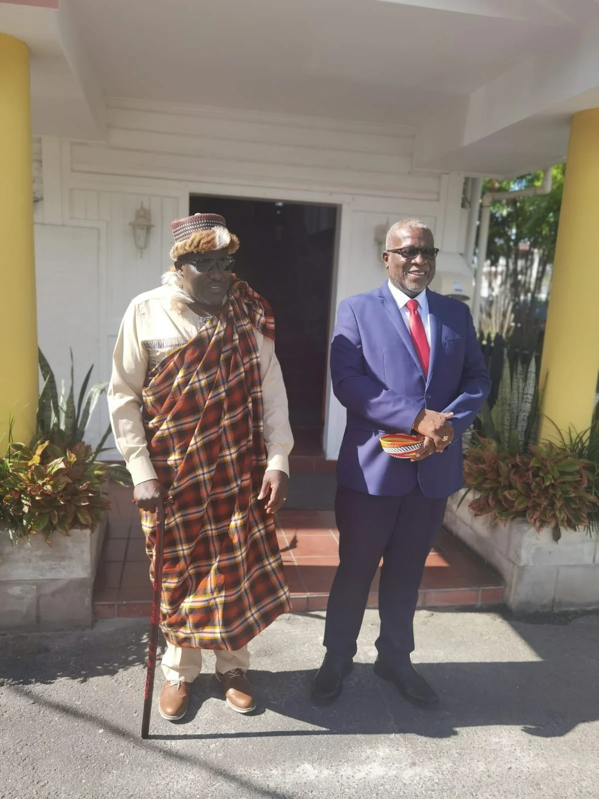 Two men standing outside a building, one dressed in traditional Maasai attire with a plaid shuka and cane, and the other in a dark blue suit with a red tie, smiling and holding a small striped item.