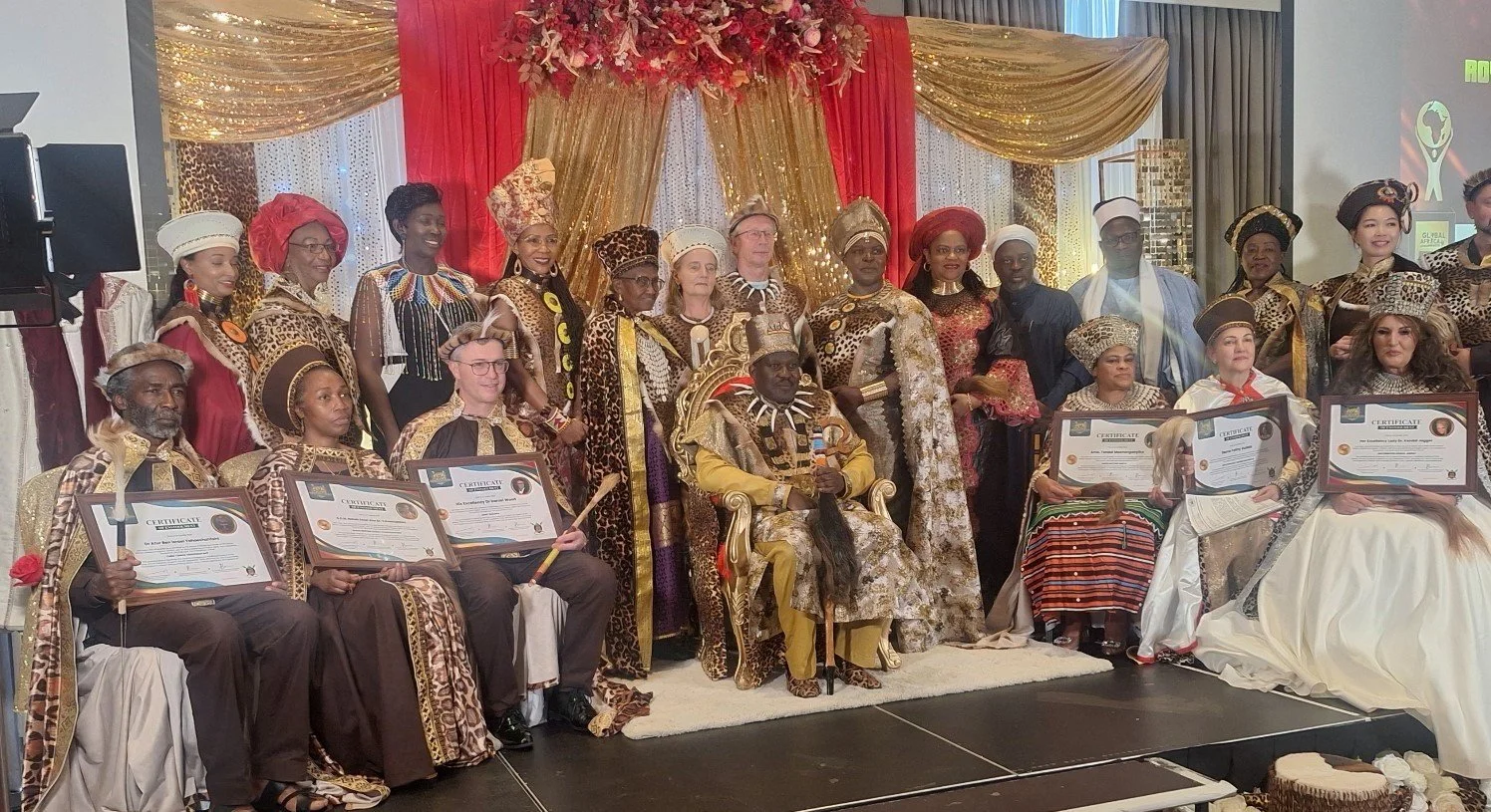 A diverse group of people dressed in traditional African attire, posing on a stage with a backdrop of red, gold, and leopard print drapes, some holding awards and certificates.