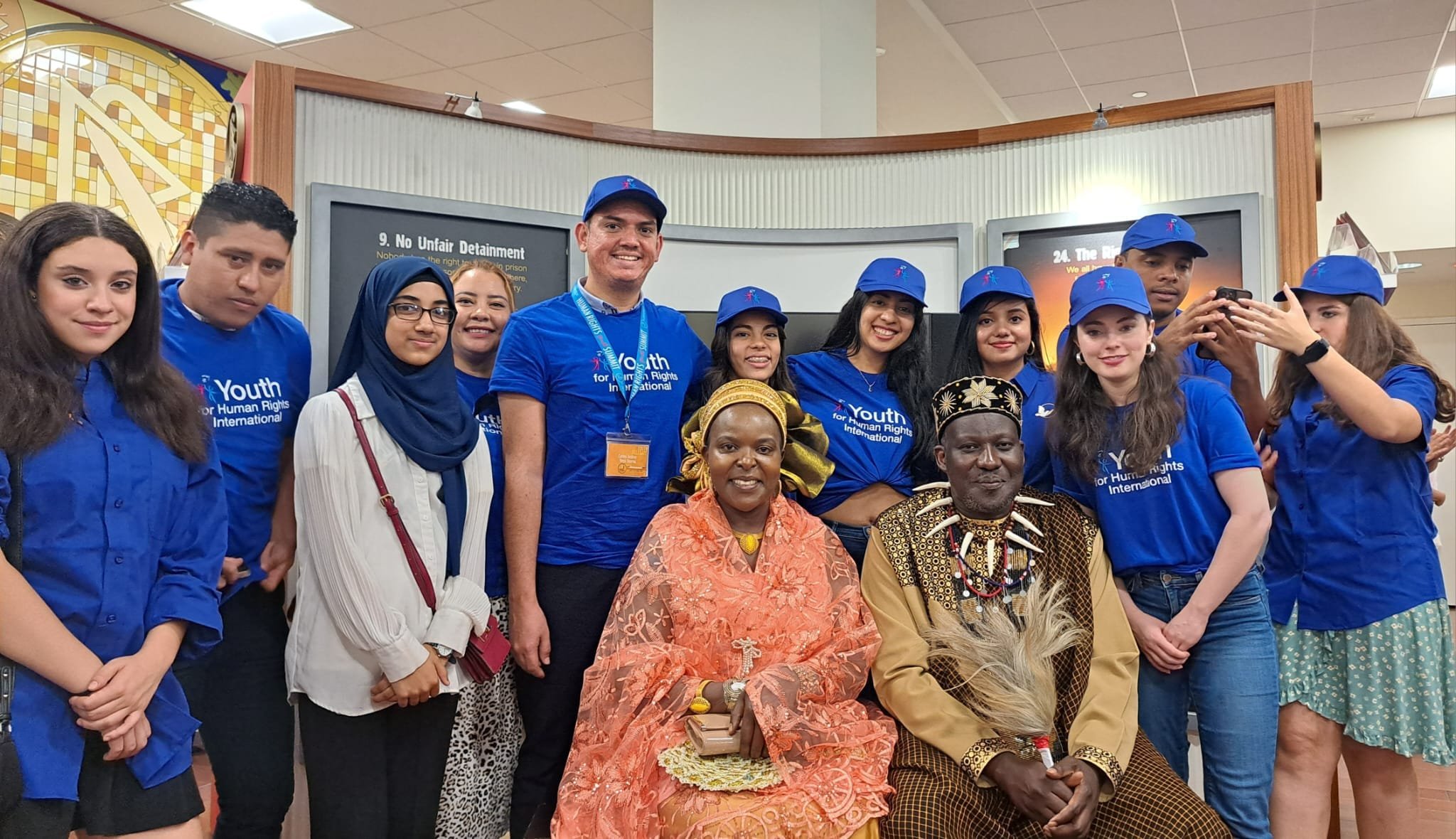 Group of young people and two adults in traditional African attire posing for a photo at an event supporting human rights. The young people are wearing blue shirts and caps with the logo of Youth for Human Rights International.