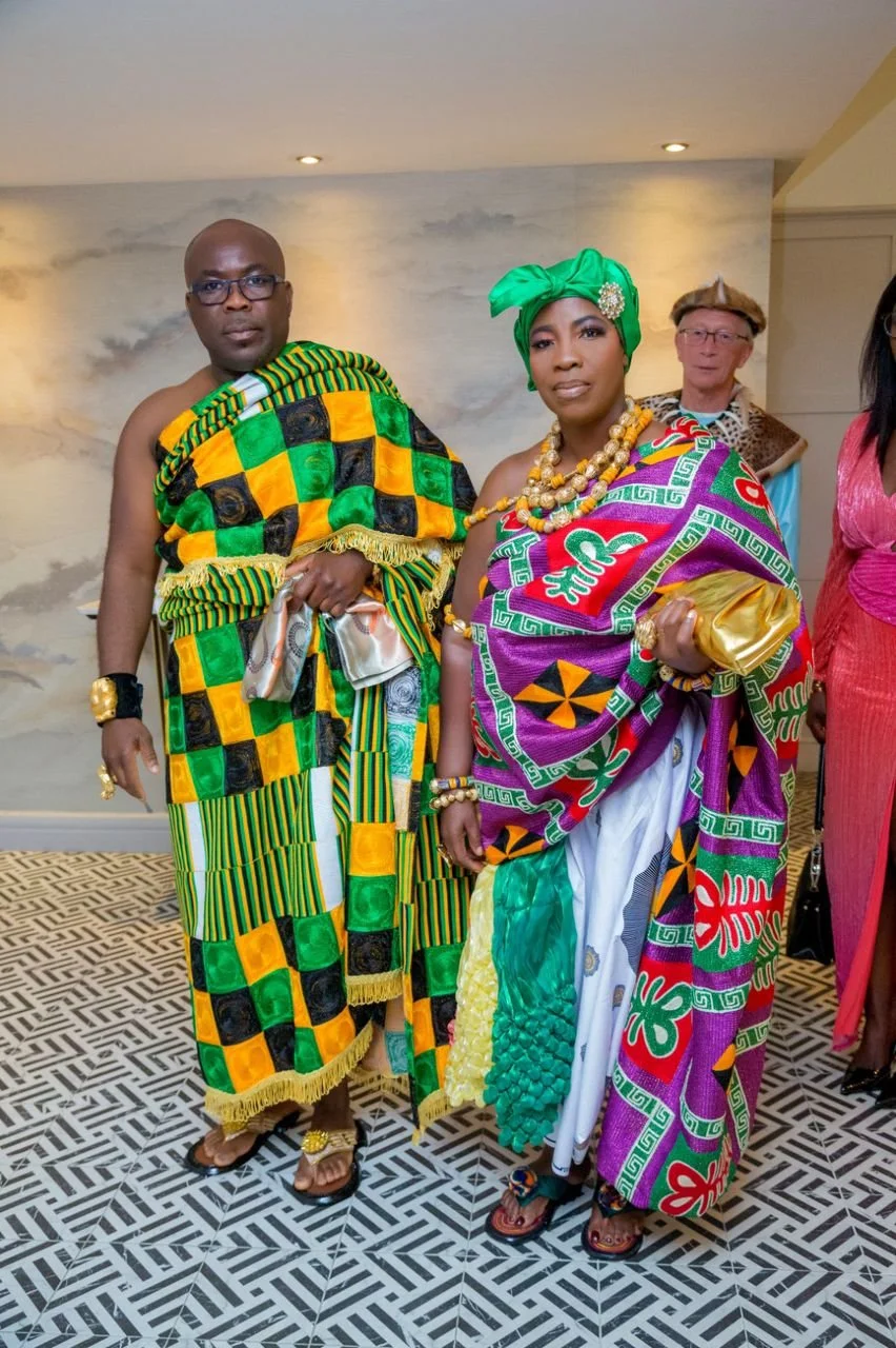 Two people dressed in vibrant traditional African attire standing indoors, with a third person in the background. The man on the left wears a kente cloth outfit, and the woman on the right wears a colorful, patterned fabric with accessories and a green headwrap.