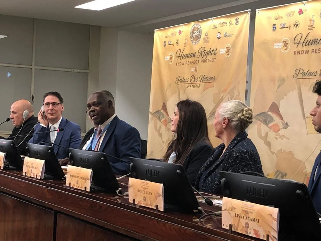 A panel of seven diverse individuals sitting at a long table with microphones and nameplates, participating in a conference about human rights at Palais Des Nations, Geneva.