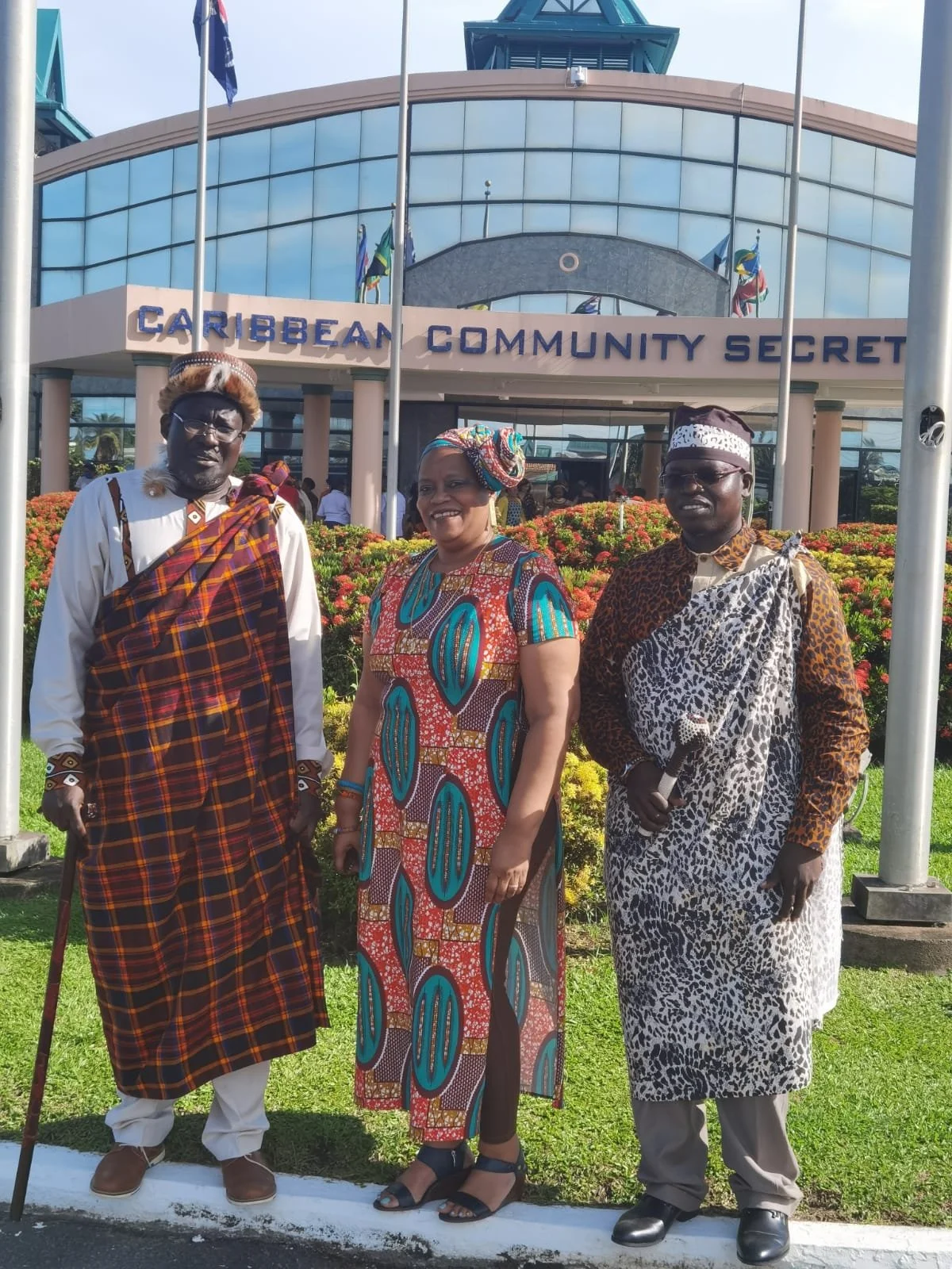Three people dressed in traditional African attire standing in front of the Caribbean Community Secretariat building.