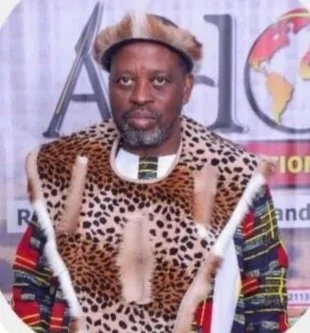 A man wearing traditional African attire with a leopard-print vest and a matching headpiece, standing in front of a backdrop with text and a globe graphic.