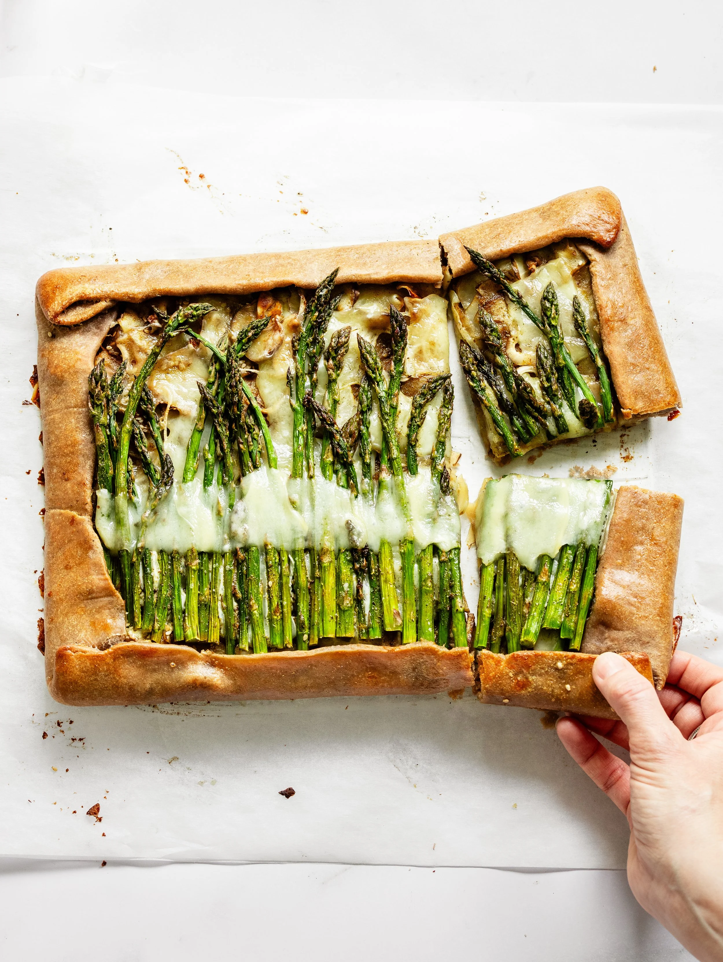 A rectangular vegetable tart with a crust, filled with green asparagus spears and melted cheese, on white parchment paper, with a person's hand holding one corner.