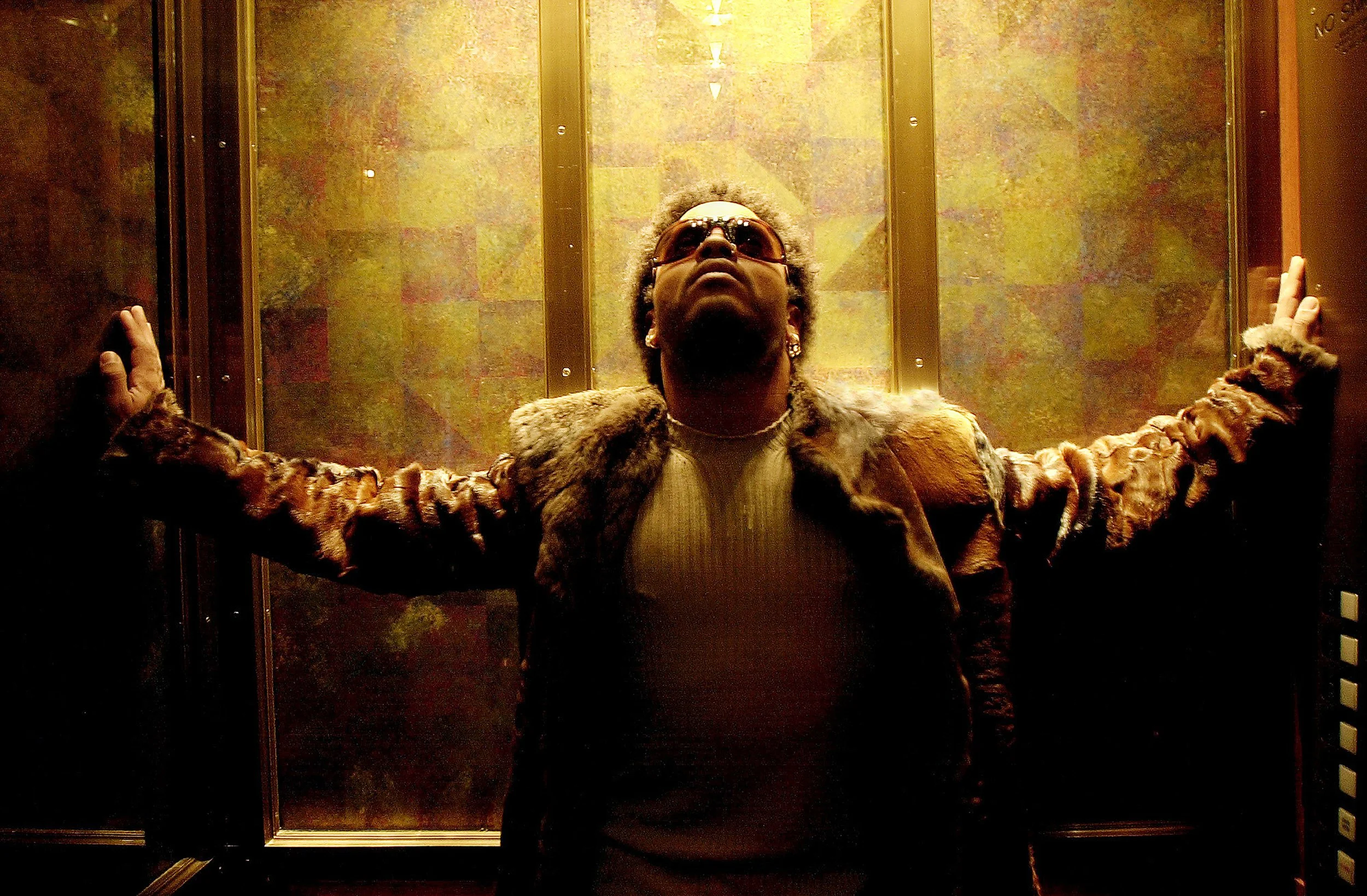A person standing with arms outstretched in an elevator, wearing sunglasses, a fur-lined jacket, and a turtleneck, with a textured wall behind them.
