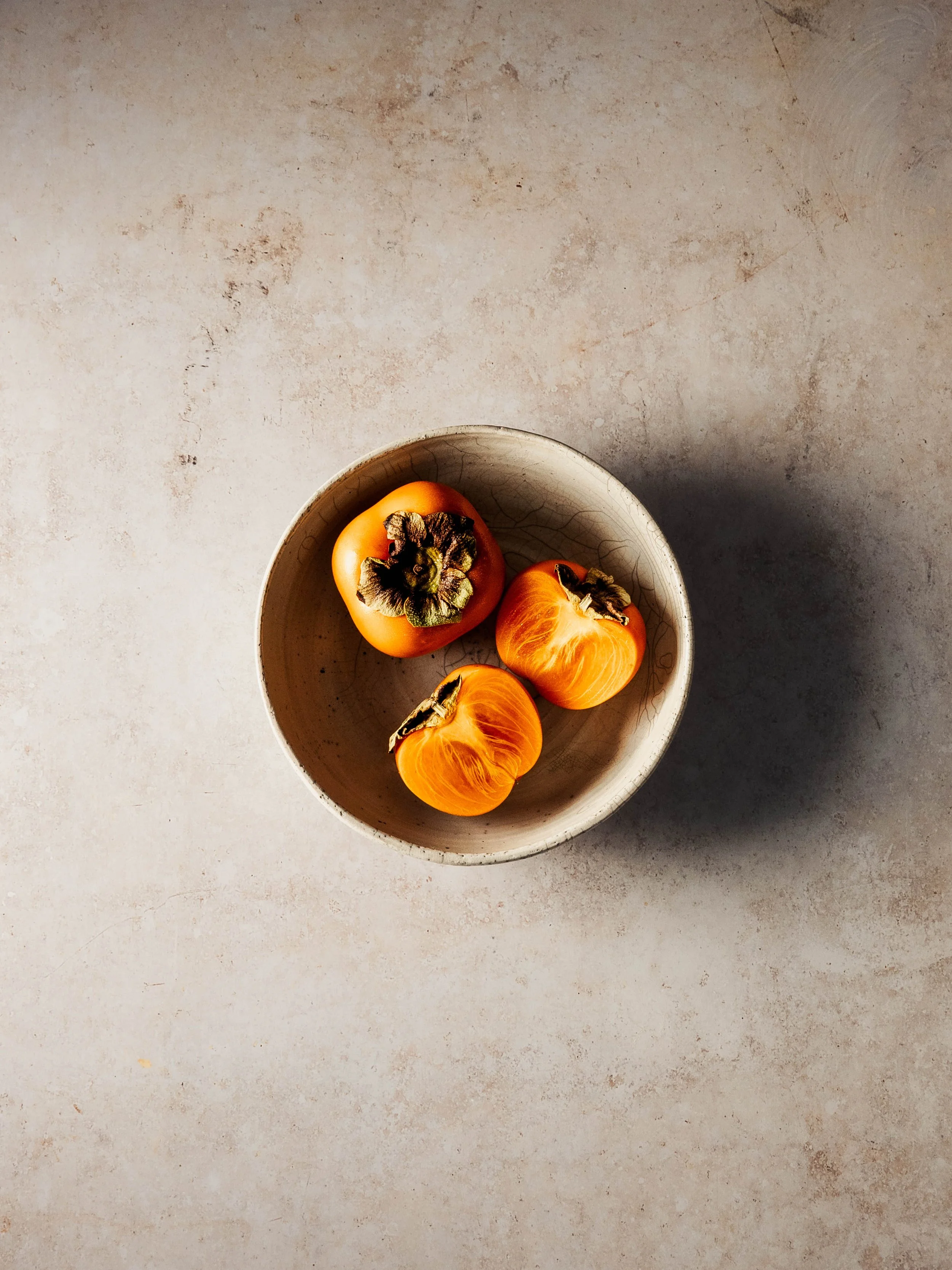 Three persimmons in a beige bowl on a textured beige surface.