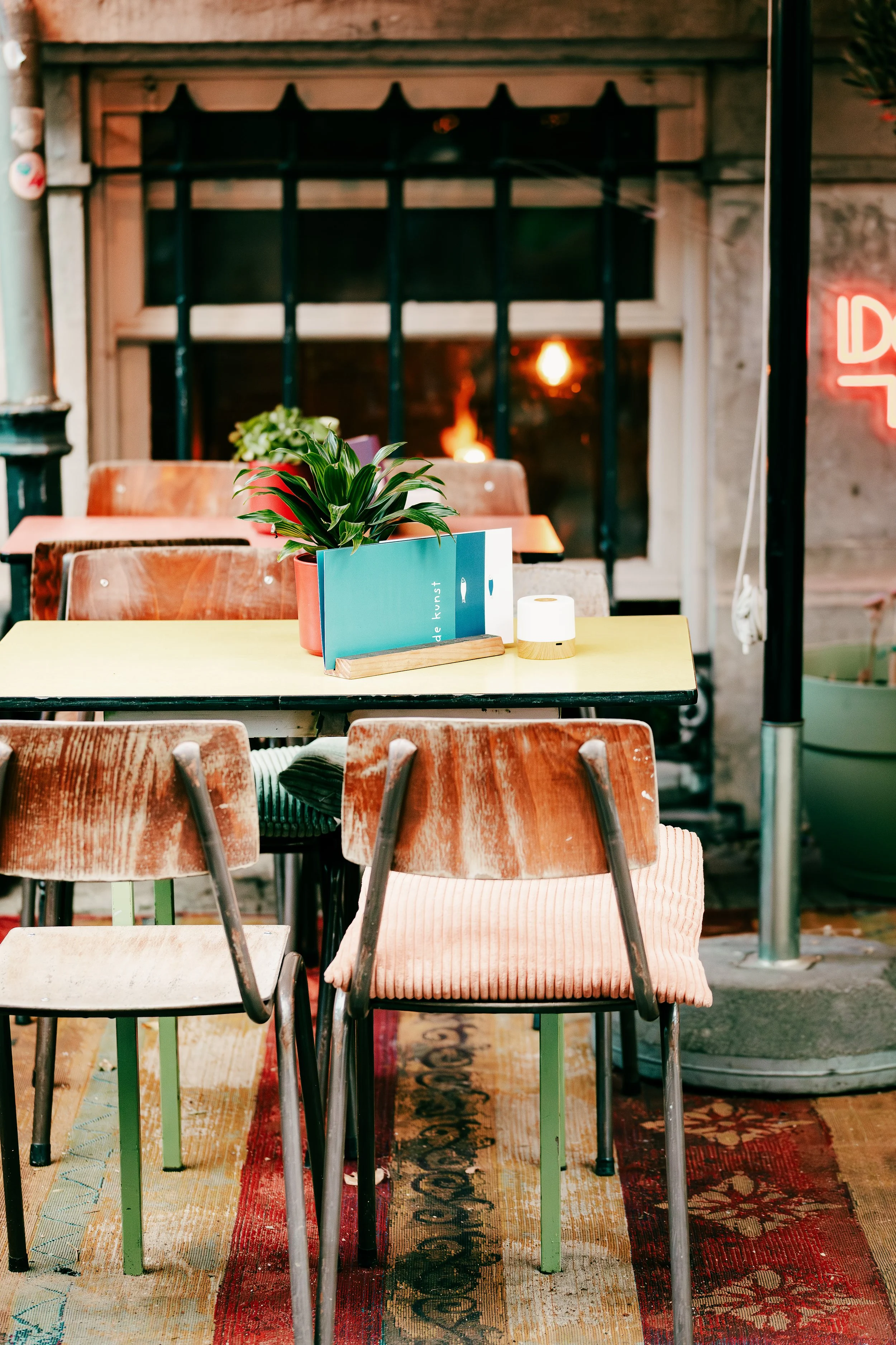 Interior of a cozy cafe with vintage wooden chairs, a table with a potted plant, a menu, and a roll of paper towels, illuminated by warm lighting.