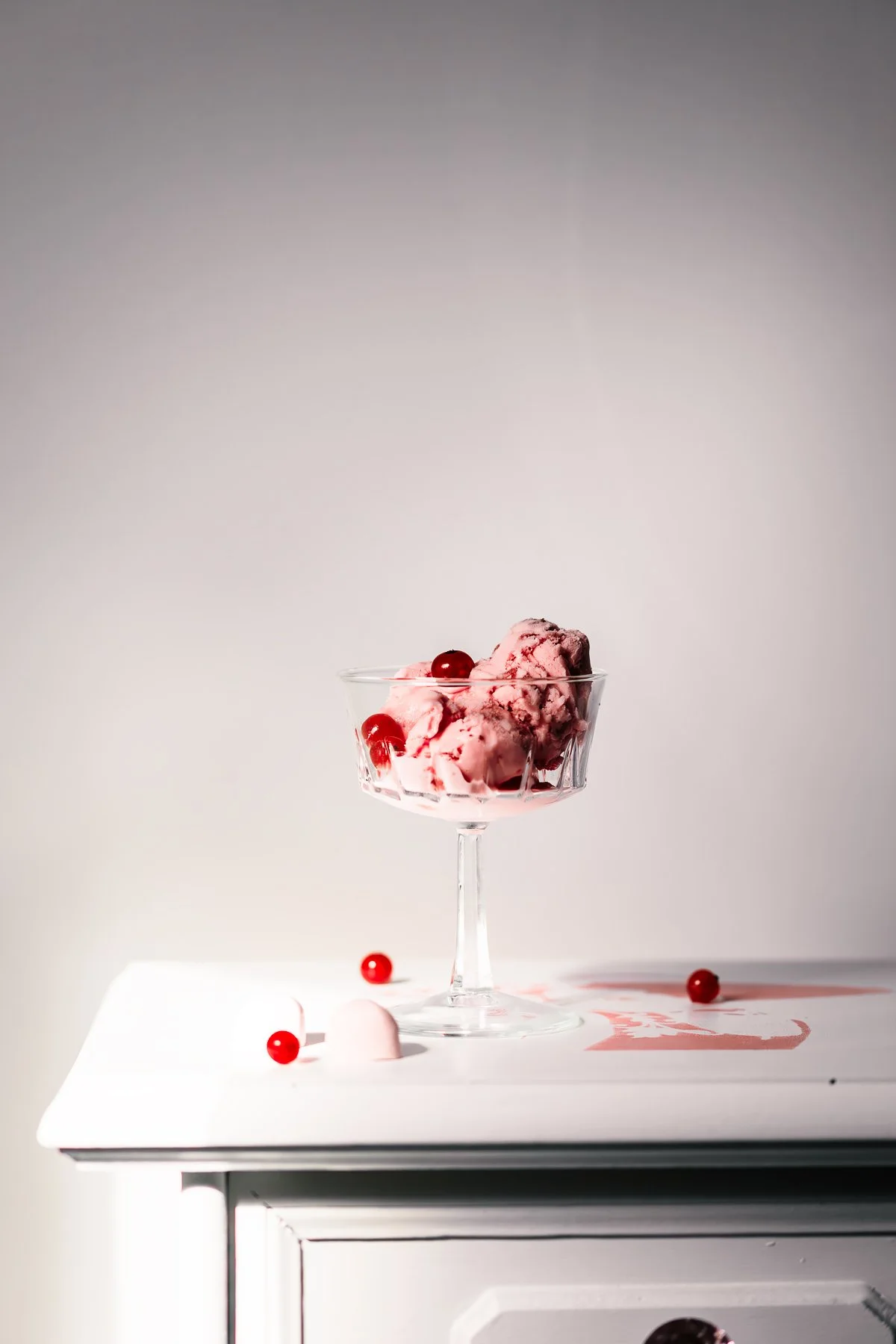 A glass bowl of pink ice cream with red berries on top, placed on a white surface with a few scattered berries and a pink design.