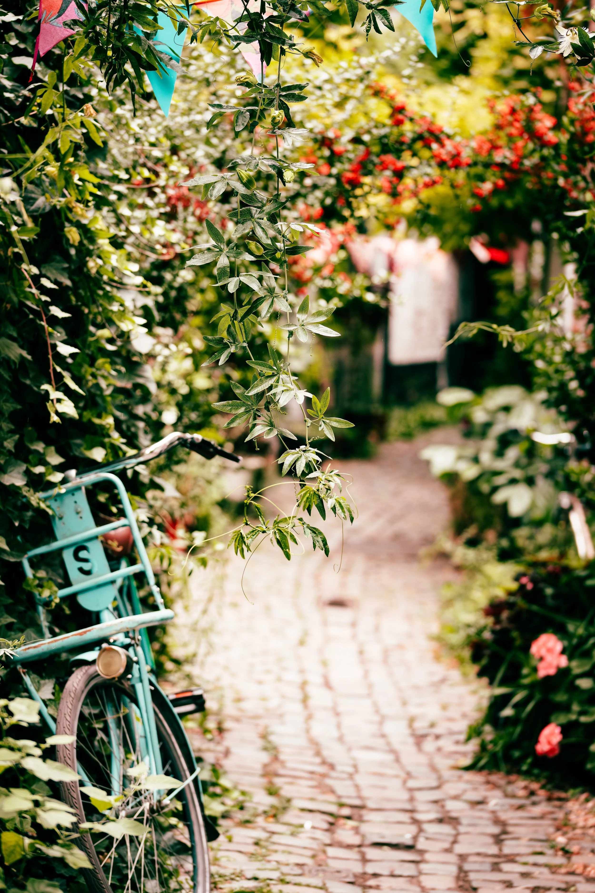 A tranquil garden pathway made of cobblestones, surrounded by lush green foliage, colorful flowers, and a vintage teal bicycle leaning against a bush, with pink and blue decorative flags hanging overhead.