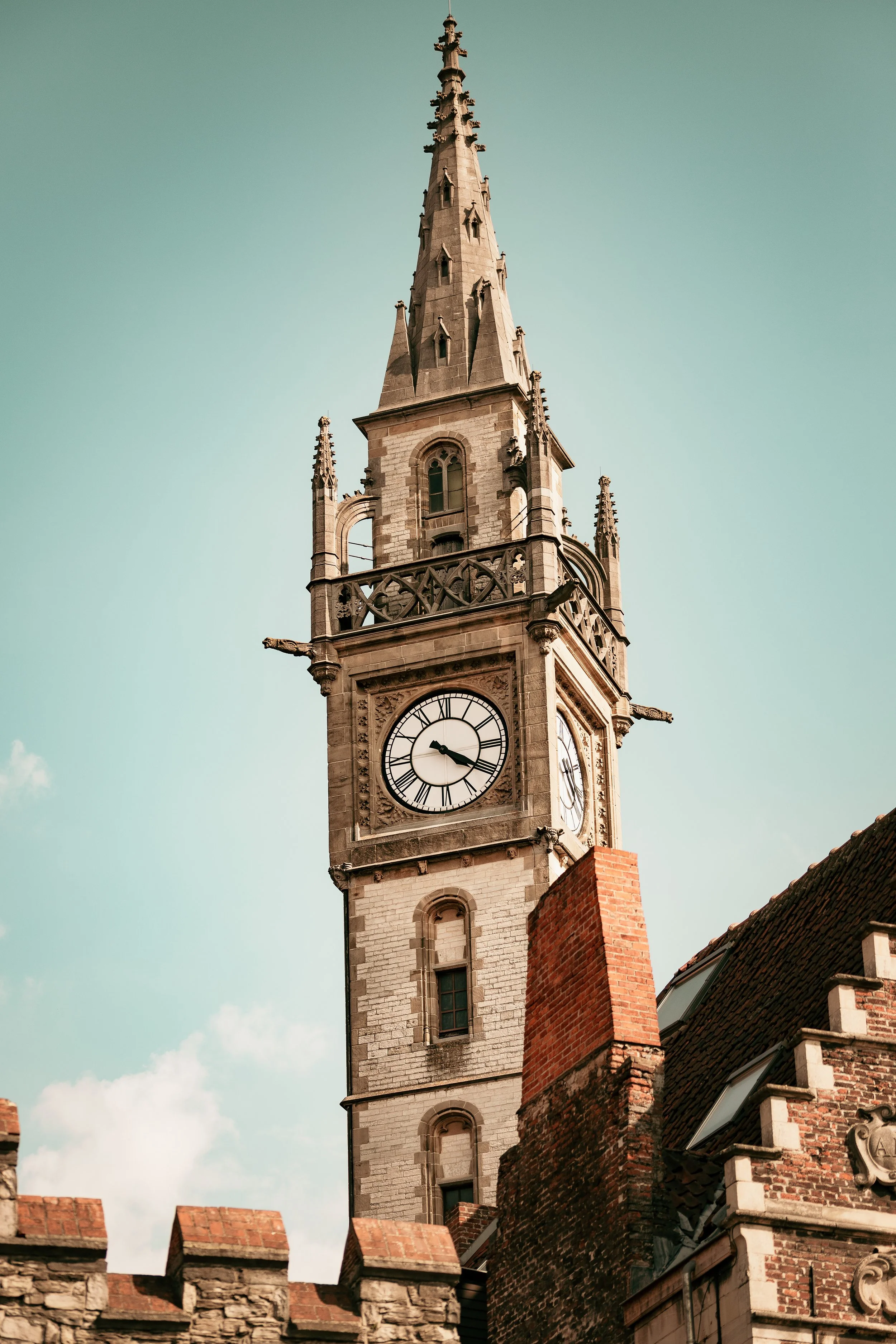A historic clock tower with a spire, surrounded by buildings with brick and stone walls, under a clear sky.
