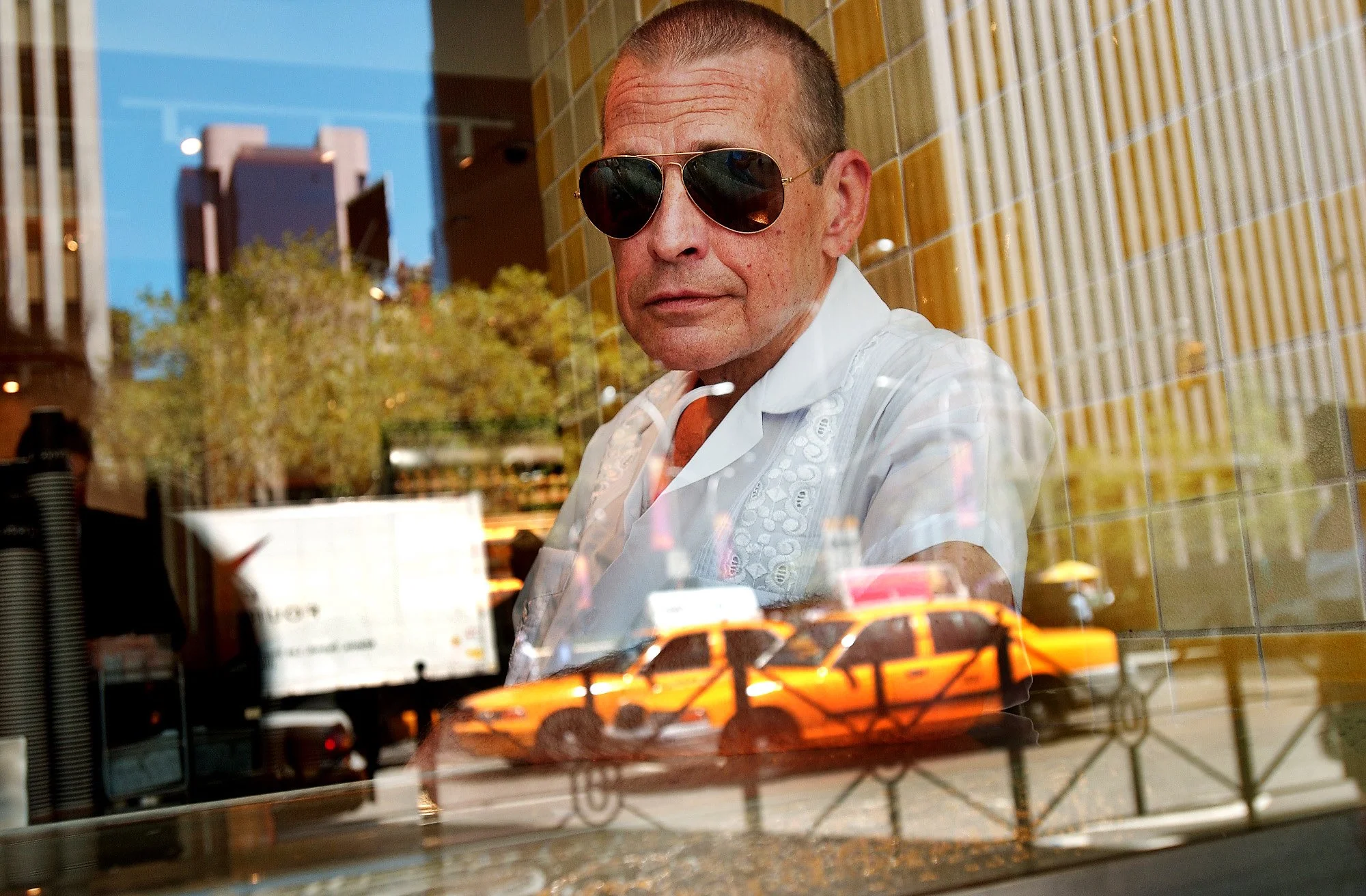 A man wearing sunglasses and a white shirt is seen through a glass window. Reflection of orange taxis and cityscape are visible on the glass.