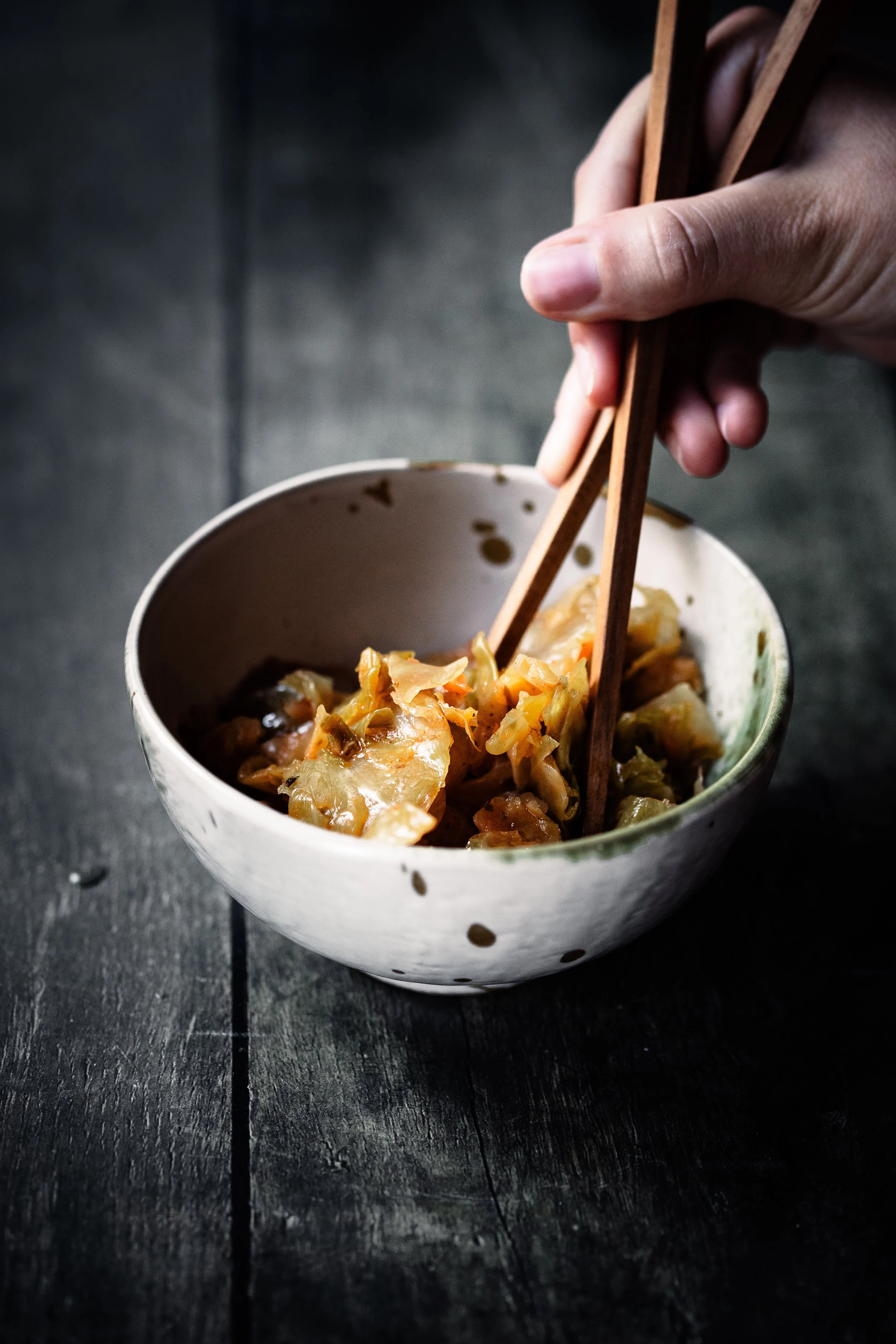 A person's hand holding wooden chopsticks over a bowl of Asian-style stir-fry with vegetables, placed on a dark wooden surface.