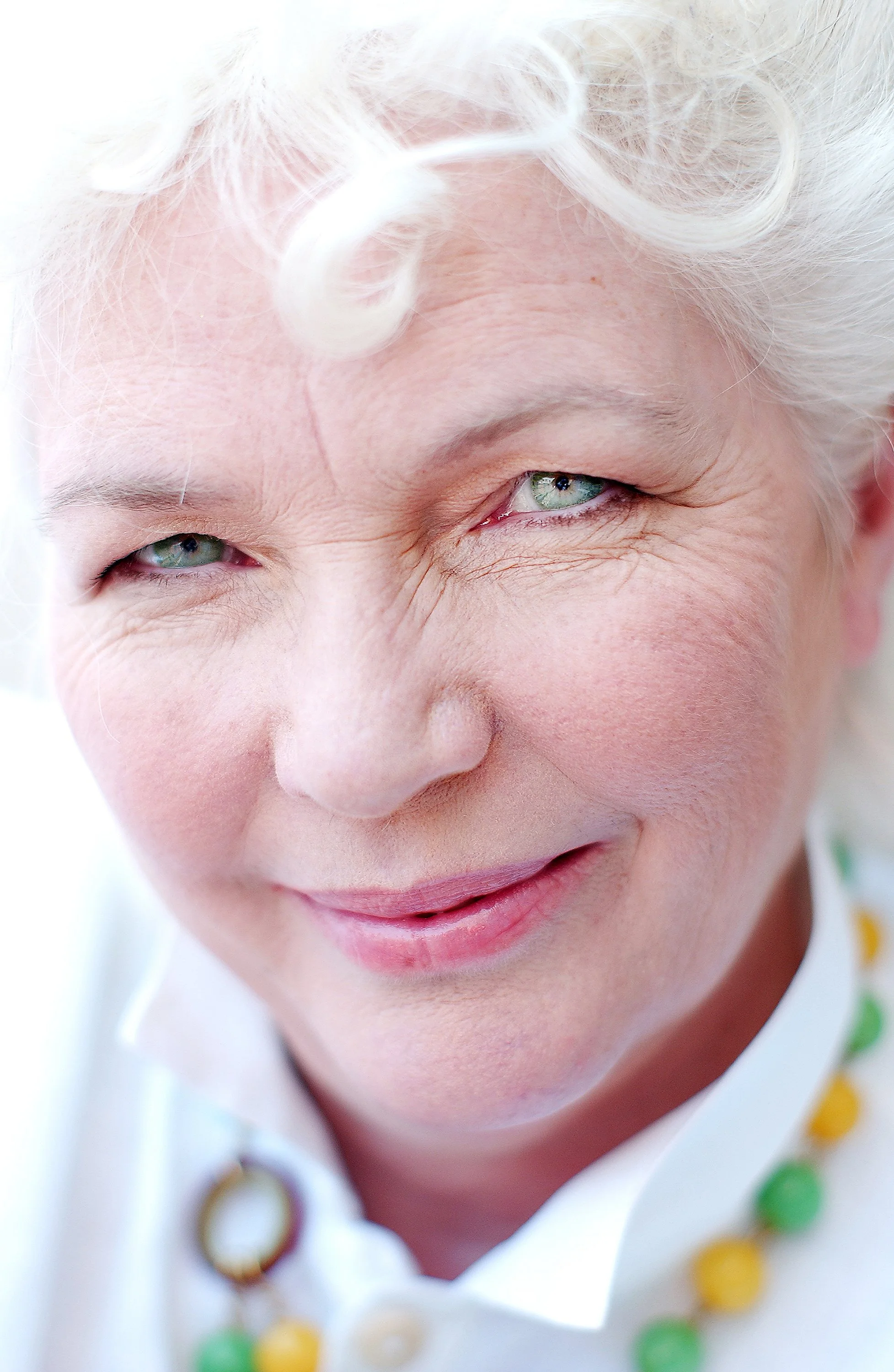 Close-up of an elderly woman with white hair, smiling with a playful expression, wearing a colorful beaded necklace.
