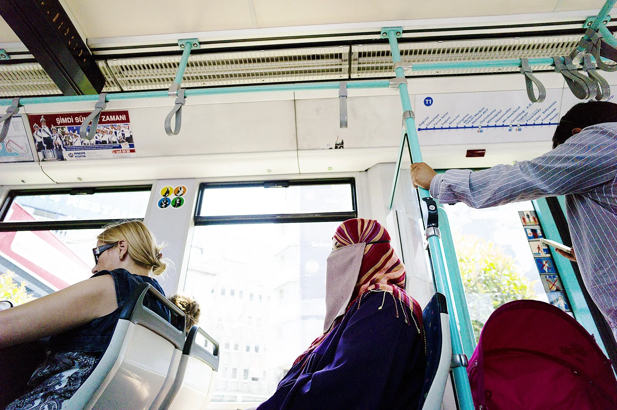 The interior of a public bus with passengers seated and standing, holding onto handrails. The bus has large windows, and some advertisements and a route map are visible.
