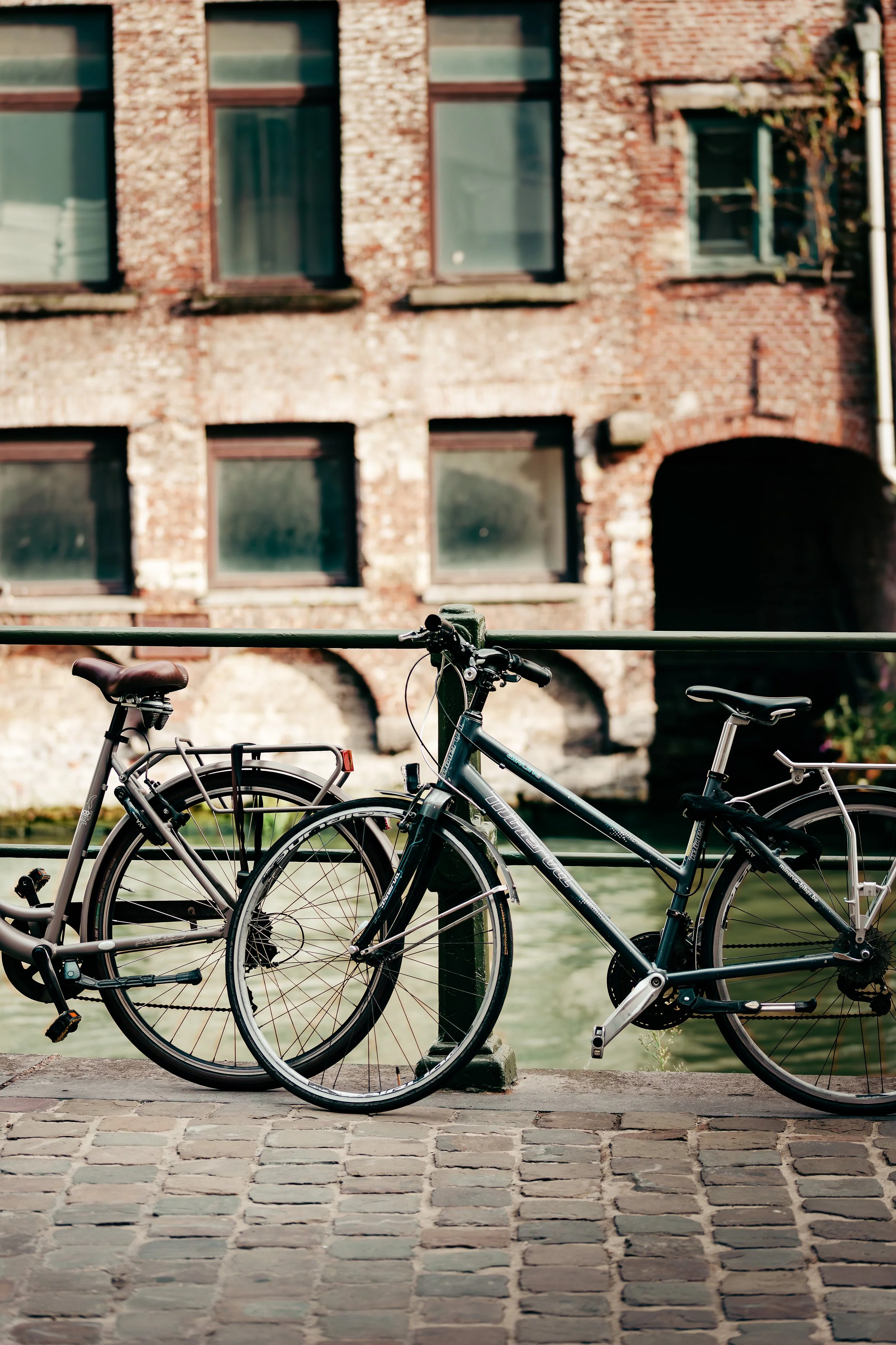 Two bicycles parked near a canal with brick buildings in the background.