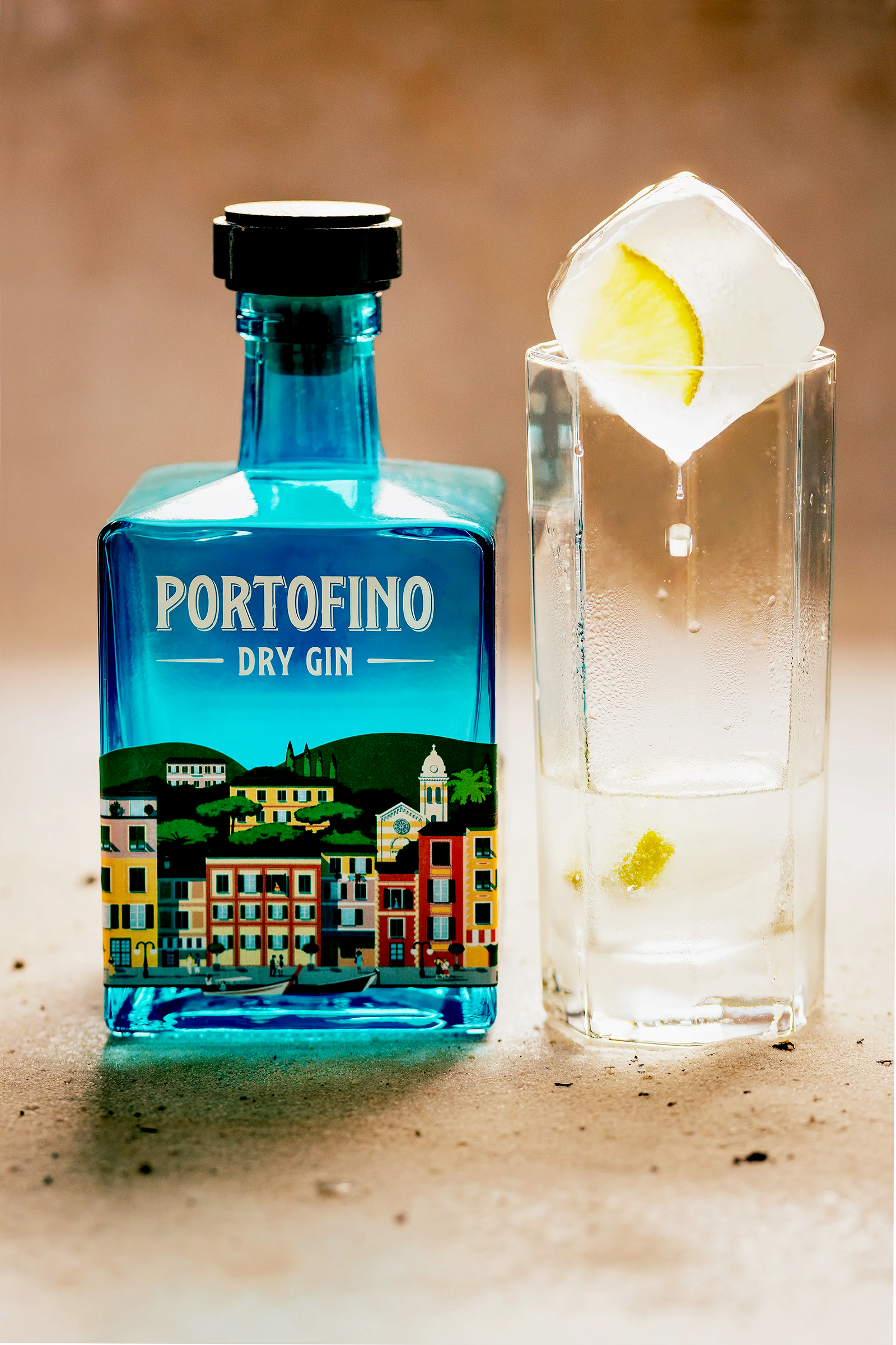A blue bottle of Portofino Dry Gin with a colorful illustration of a town with buildings and hills on the label, placed next to a clear glass with a large ice cube and a lemon wedge.