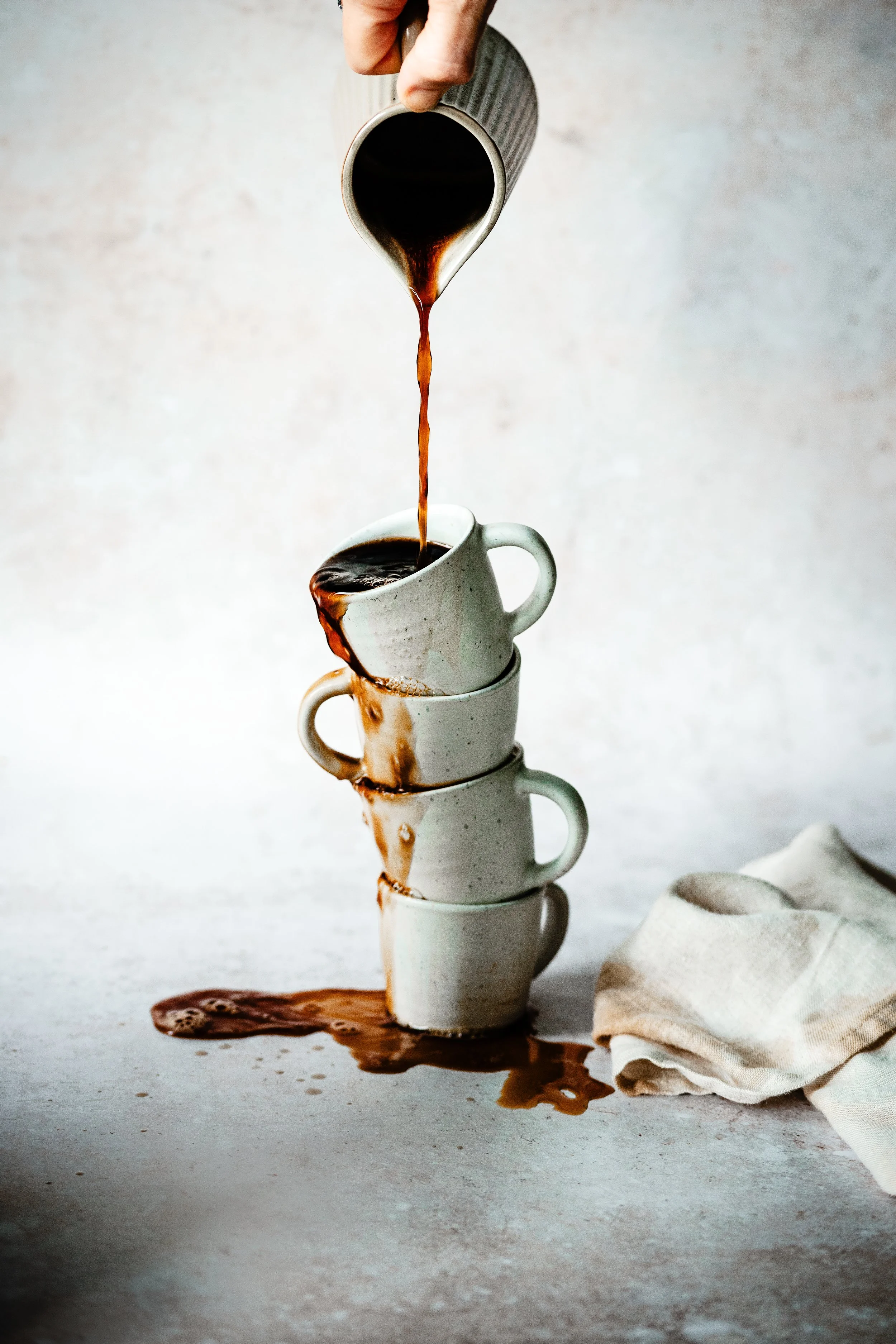 A hand pouring coffee from a mug into a stack of three white mugs on a table. Coffee spills over the table, and a cloth is placed beside the mugs.