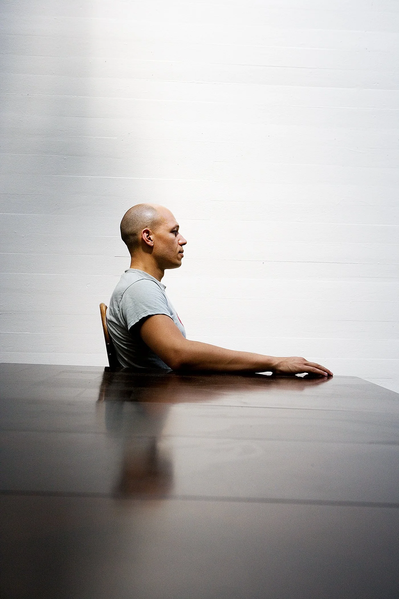 A man with a shaved head sitting at a wooden table in profile view against a plain white wall.
