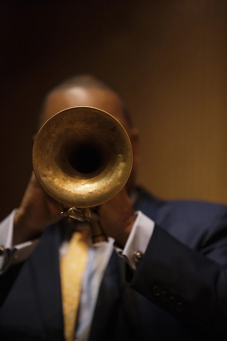 A person in a suit playing a trumpet, the camera focuses on the bell of the trumpet, obscuring the person's face.