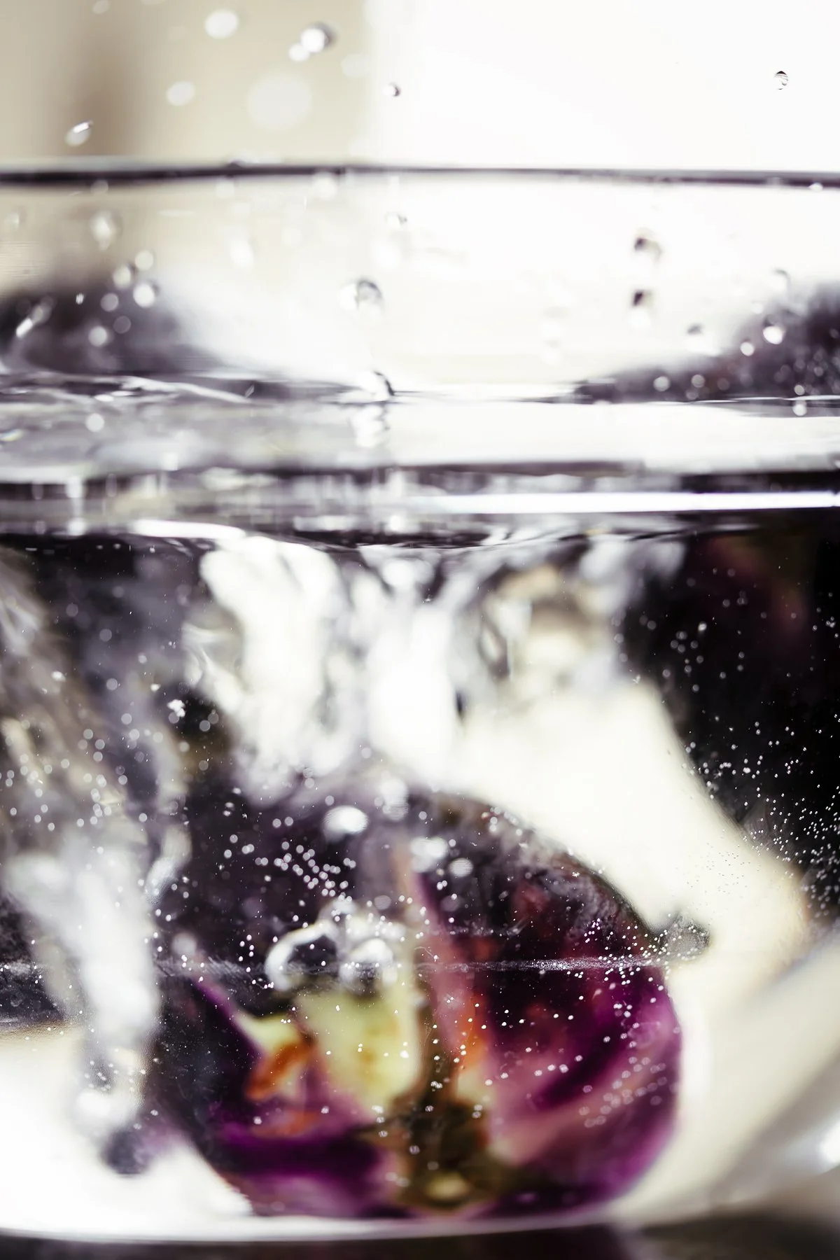 A close-up photo of a purple and white eggplant being submerged in water, creating bubbles and splashes.