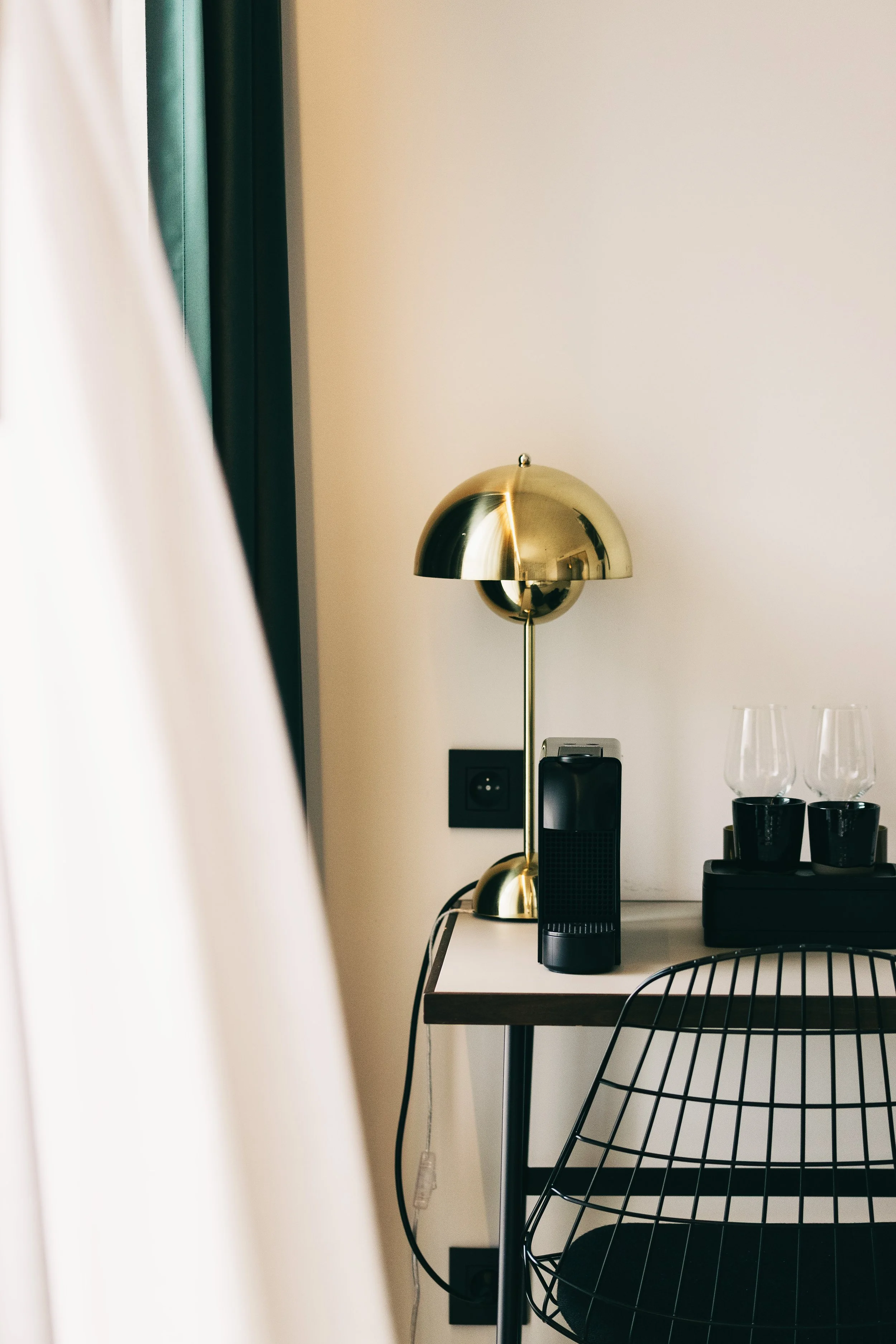A corner of a room with a small desk, a black chair, a gold desk lamp, a black air purifier or speaker, and a tray with four glassware cups on it.