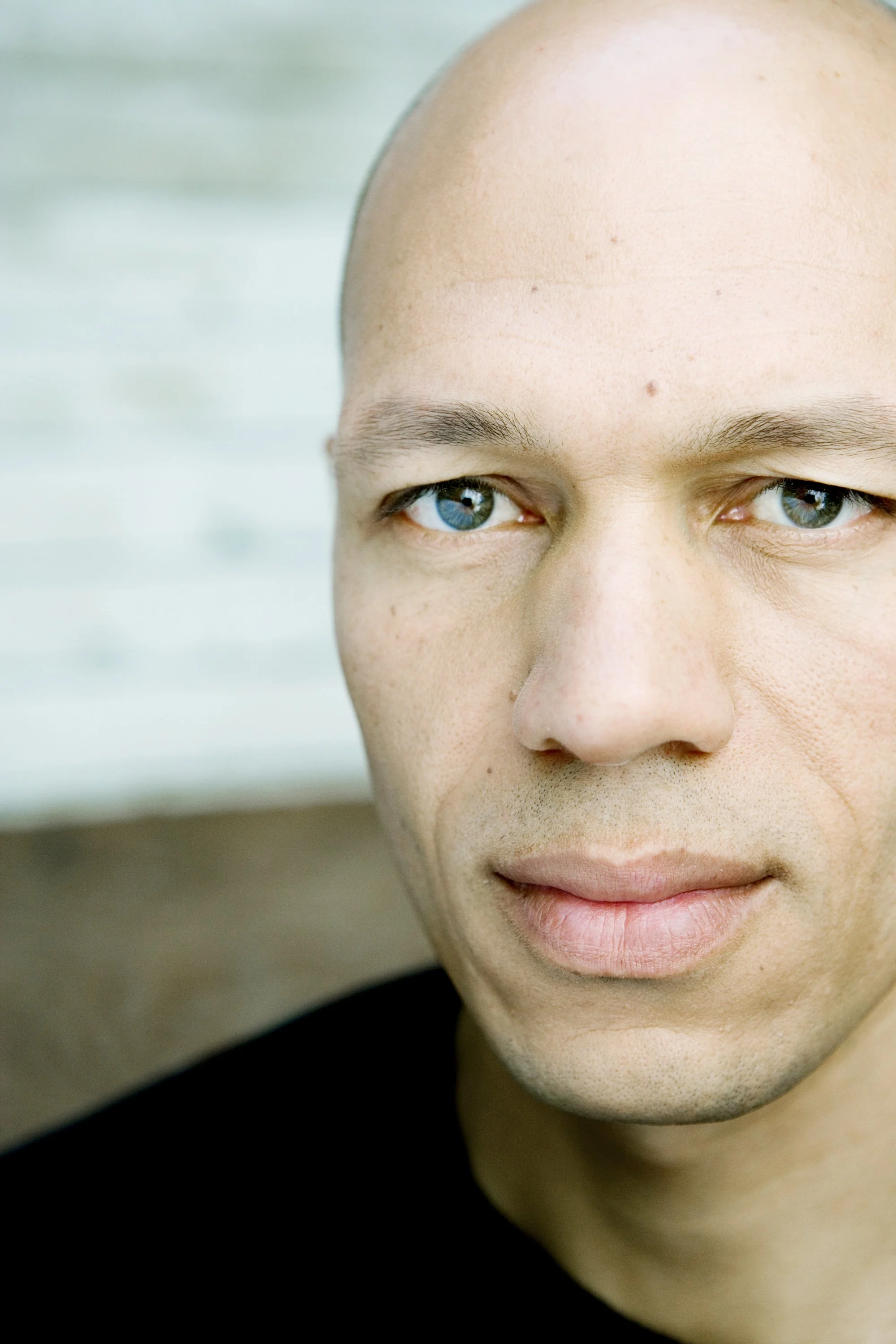 Close-up of a young man with blue eyes, light skin, and a shaved head, looking slightly to the right against a blurred background.