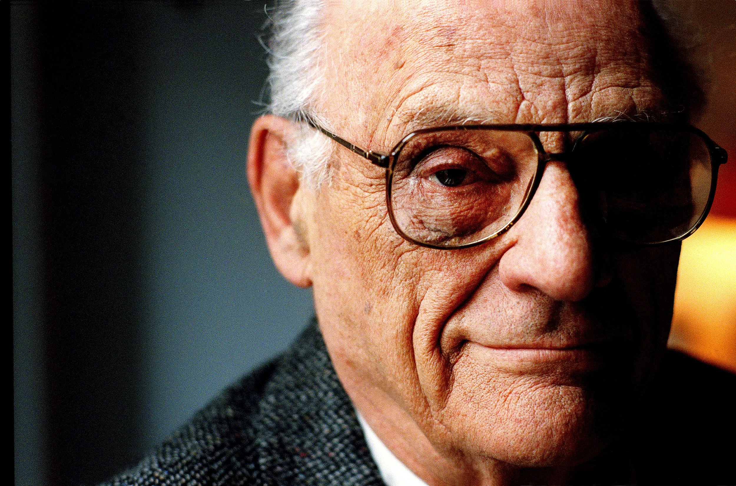 Close-up of an elderly man with white hair, wearing glasses, and covering part of his face with his hand.