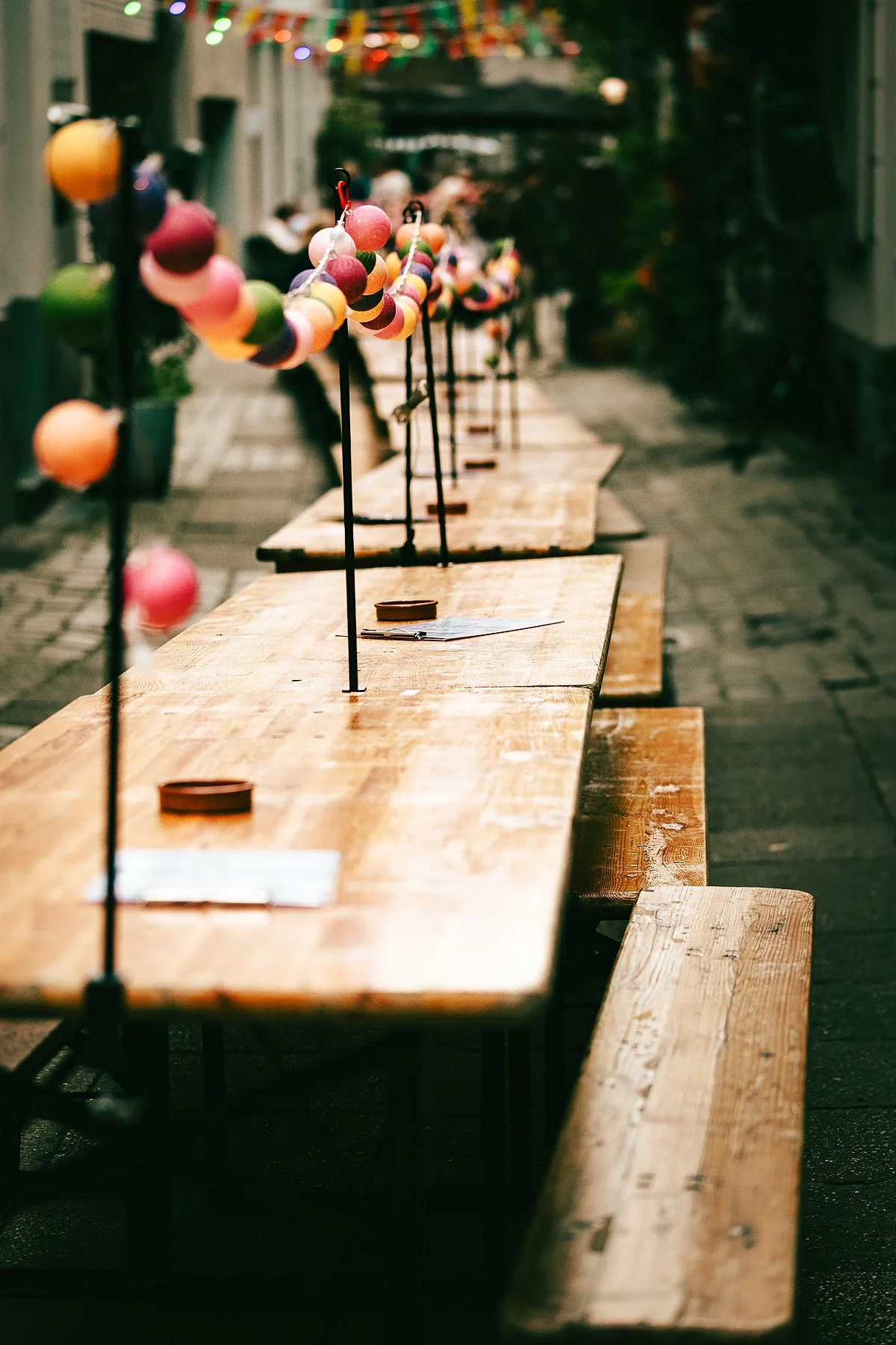 Empty outdoor wooden tables and benches with colorful string lights hanging overhead, set up for a celebration or event.