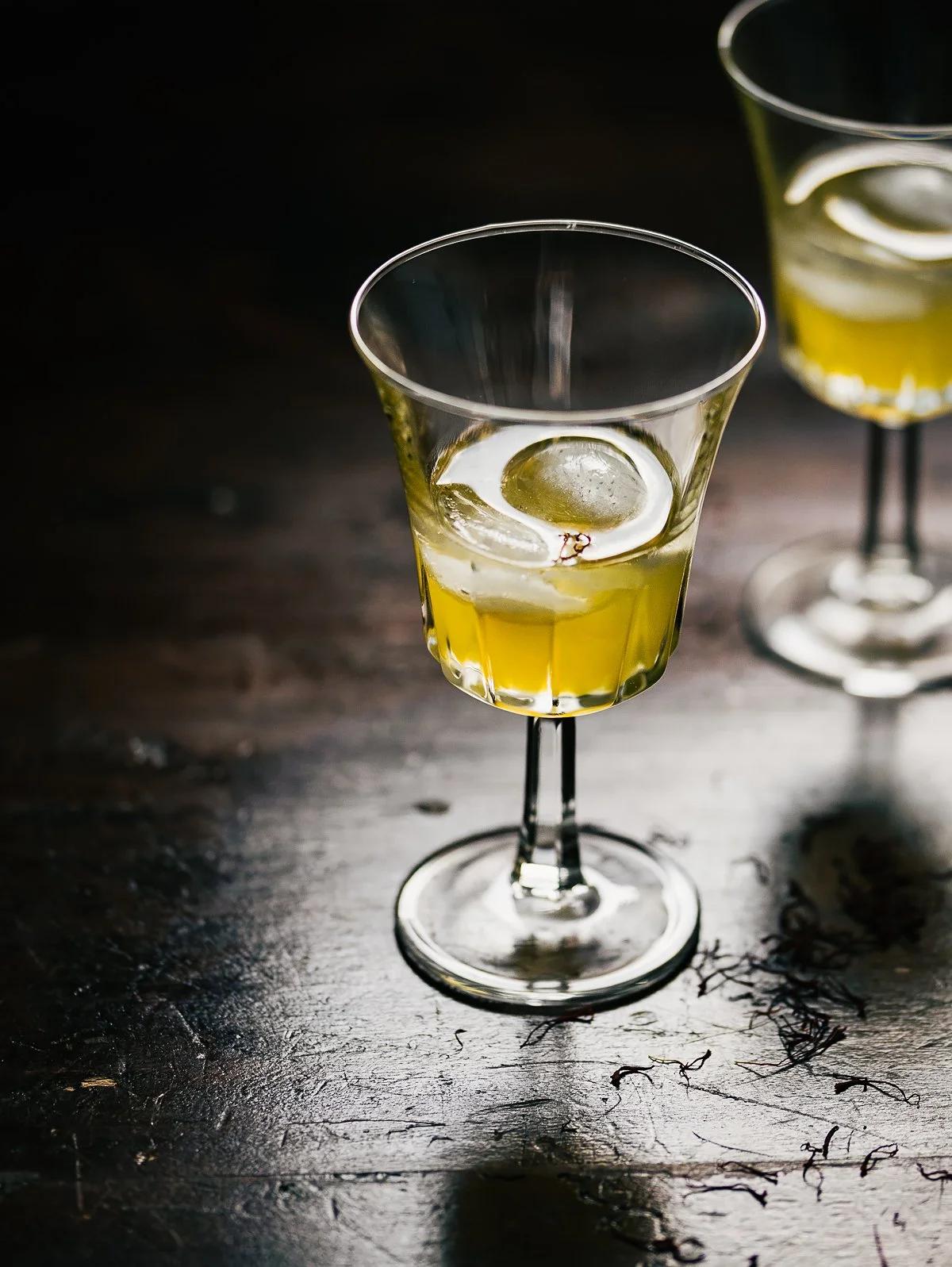 Two cocktail glasses with yellow liquid and large ice cubes on a dark wooden surface.