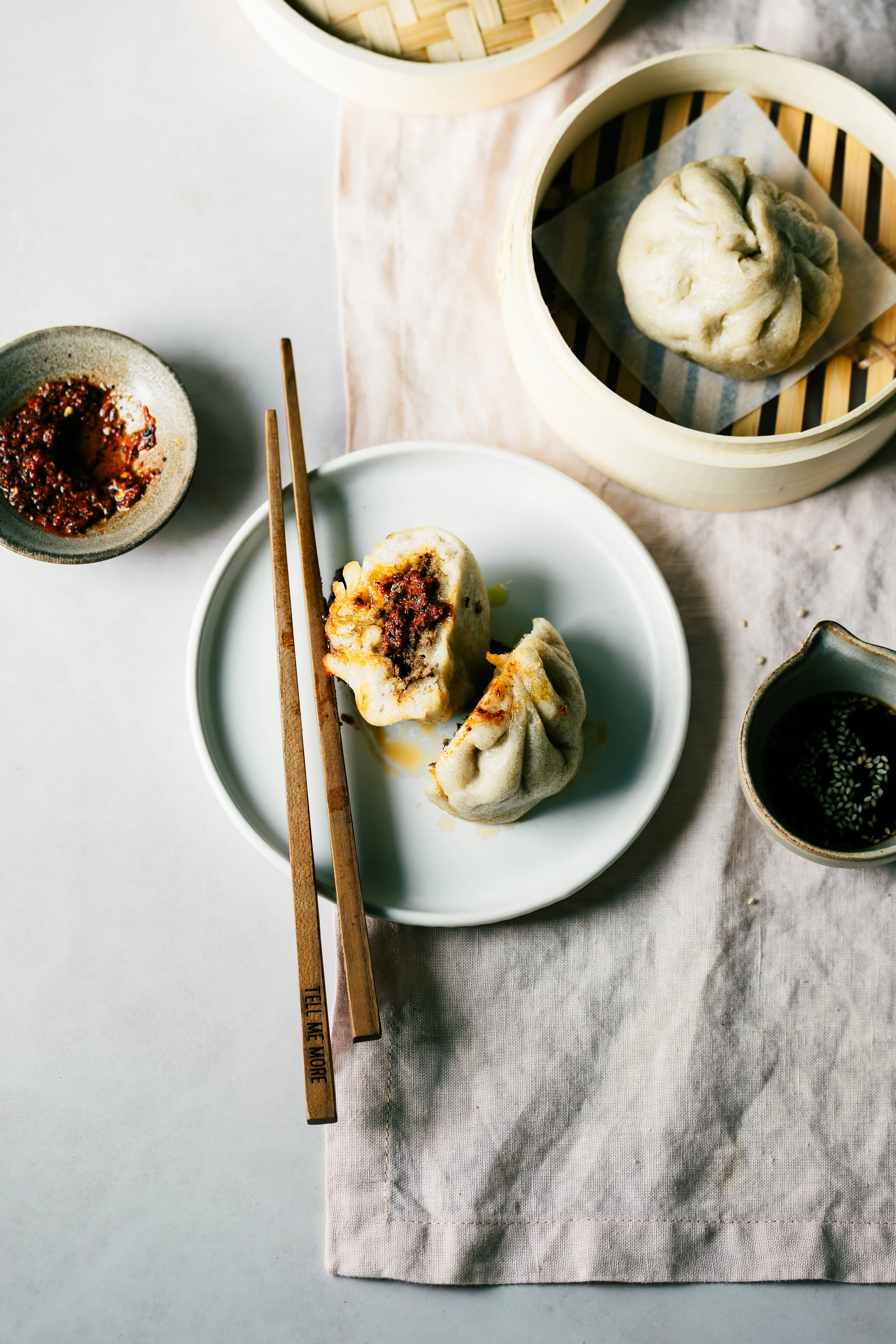 A steamed bun with filling on a white plate, chopsticks, two bamboo steamers, and small bowls of sauce on a linen tablecloth.