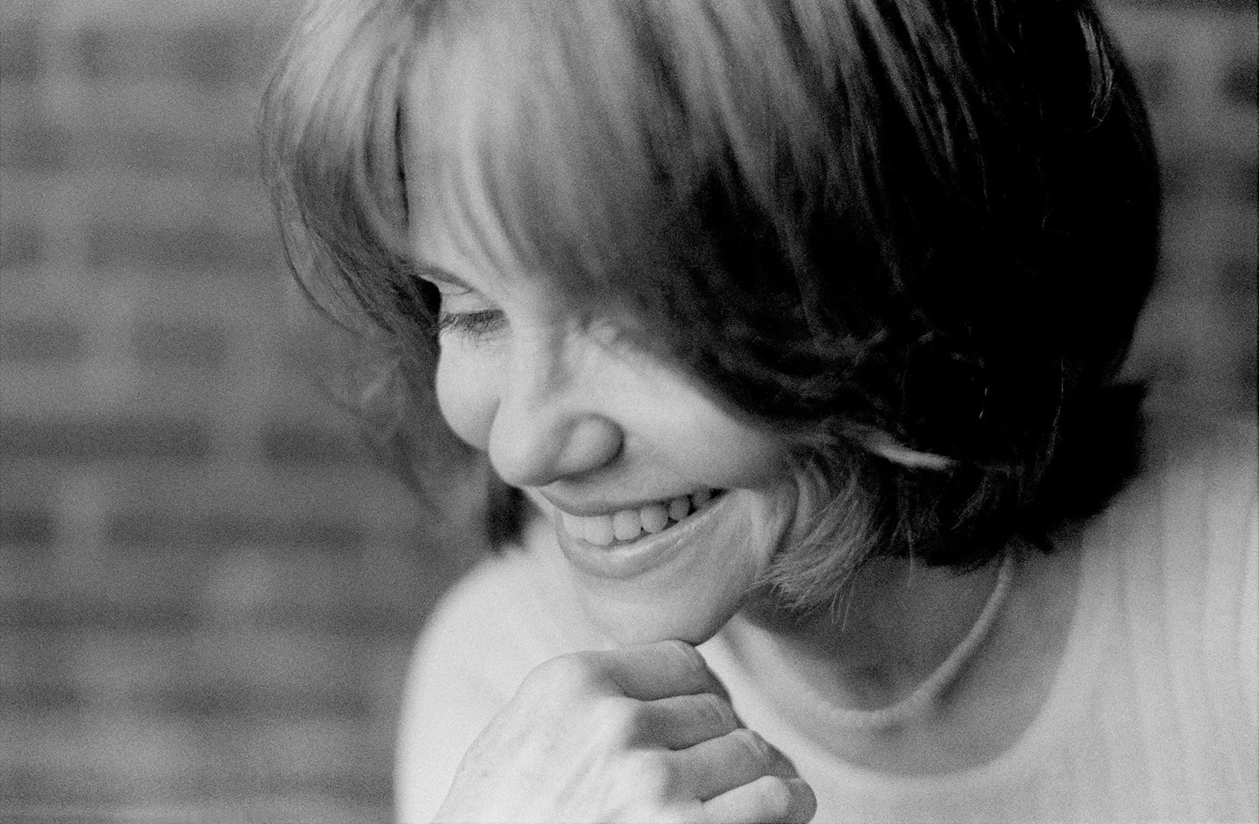Black and white close-up photo of a smiling woman with shoulder-length hair, looking downward, with a hand near her chin.