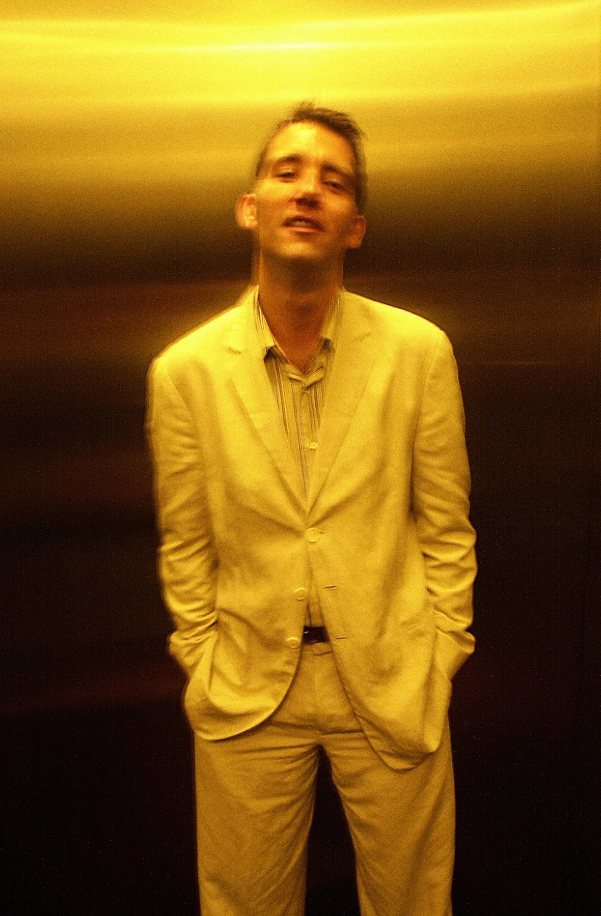A man in a beige suit and striped shirt standing in front of a yellow and black background, with a slight blur and motion effect.