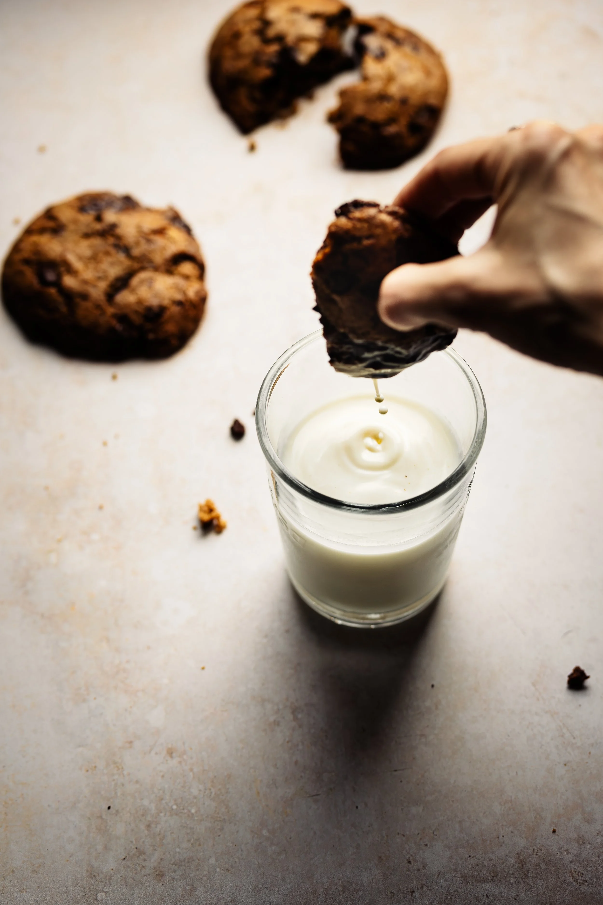 A hand holding a cookie over a glass of milk, with two more cookies in the background on a light-colored surface. The cookies are chocolate chip and appear homemade.