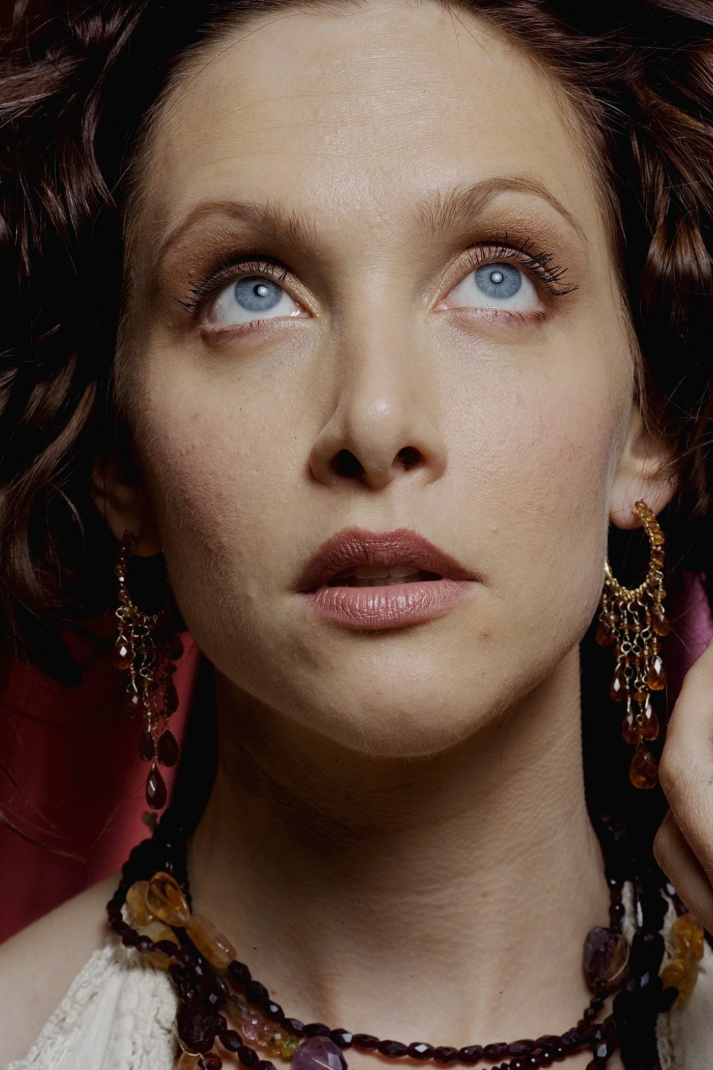 Close-up of a woman with bright blue eyes, curly dark hair, and wearing jewelry including earrings and a necklace, looking upward.