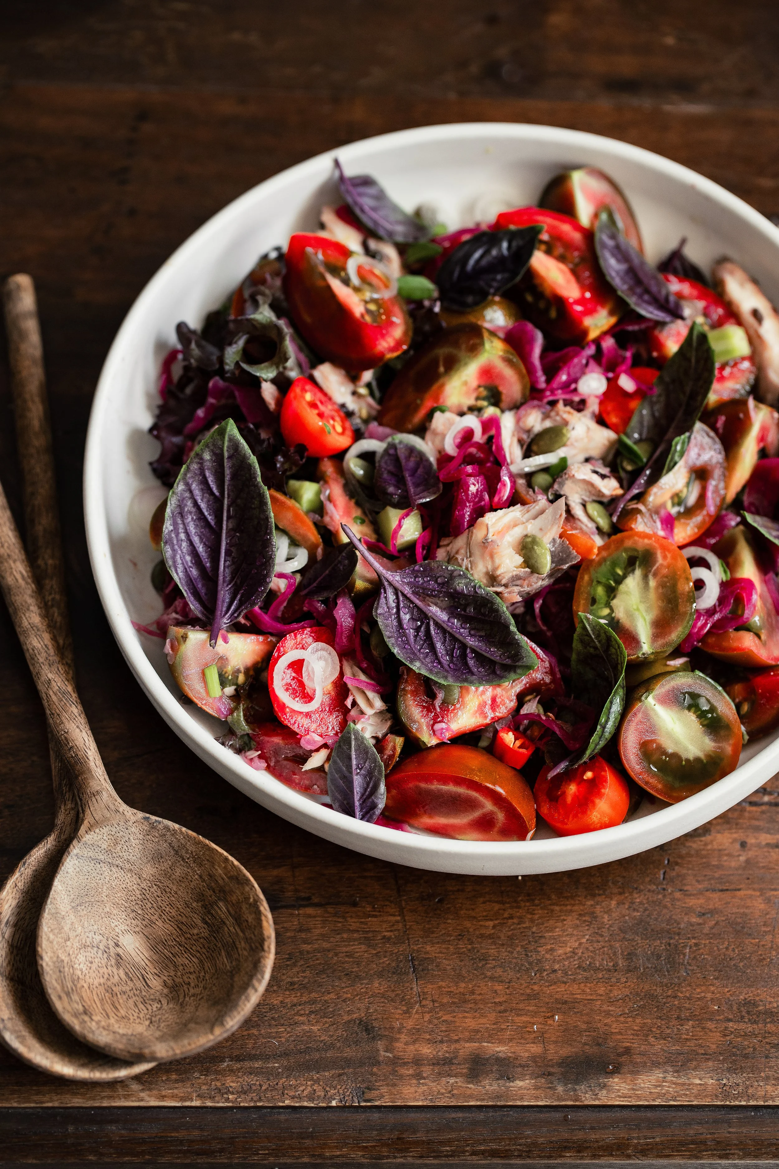 A white bowl filled with a colorful salad containing cherry tomatoes, purple basil leaves, shredded purple cabbage, chopped green onions, and pieces of chicken on a dark wooden surface. Two wooden spoons are placed next to the bowl.