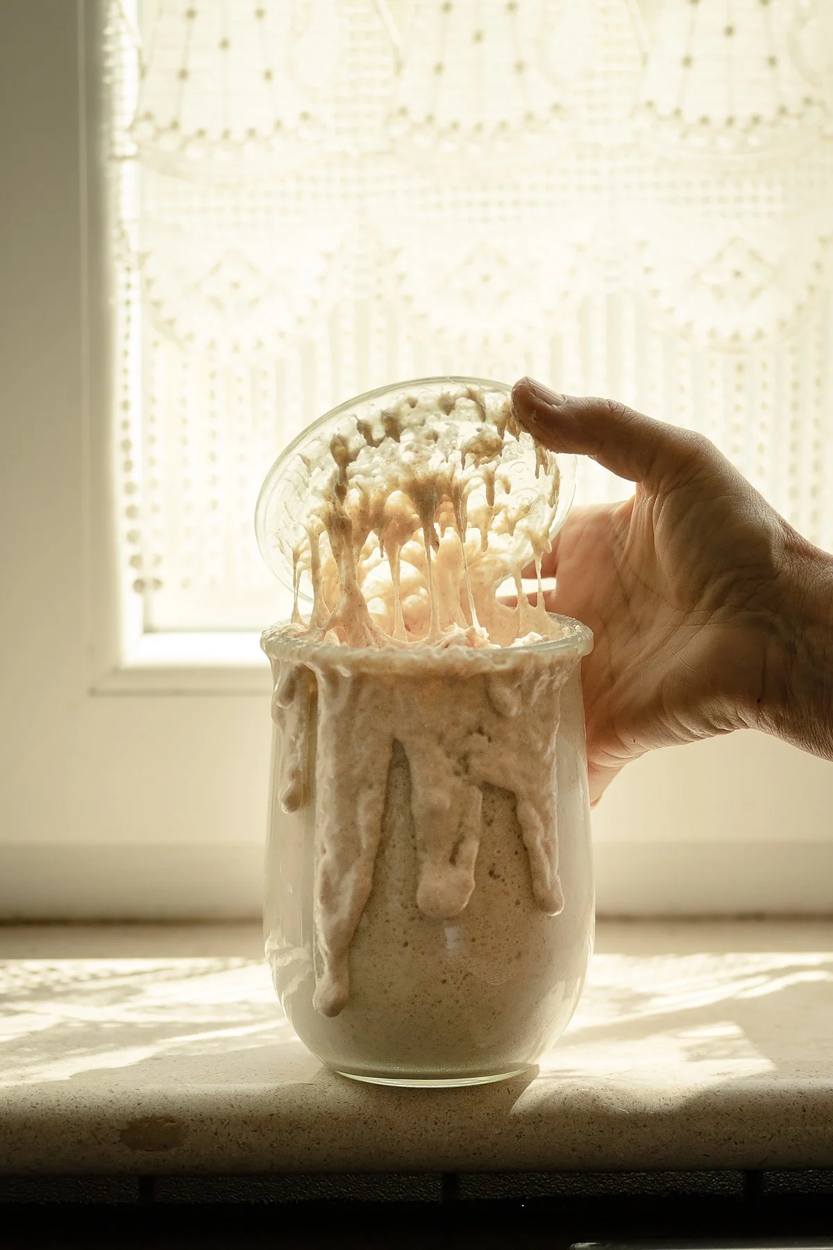 A hand lifting a spoonful of overflowing, creamy, light-colored substance from a glass jar, with a window and lace curtain in the background.