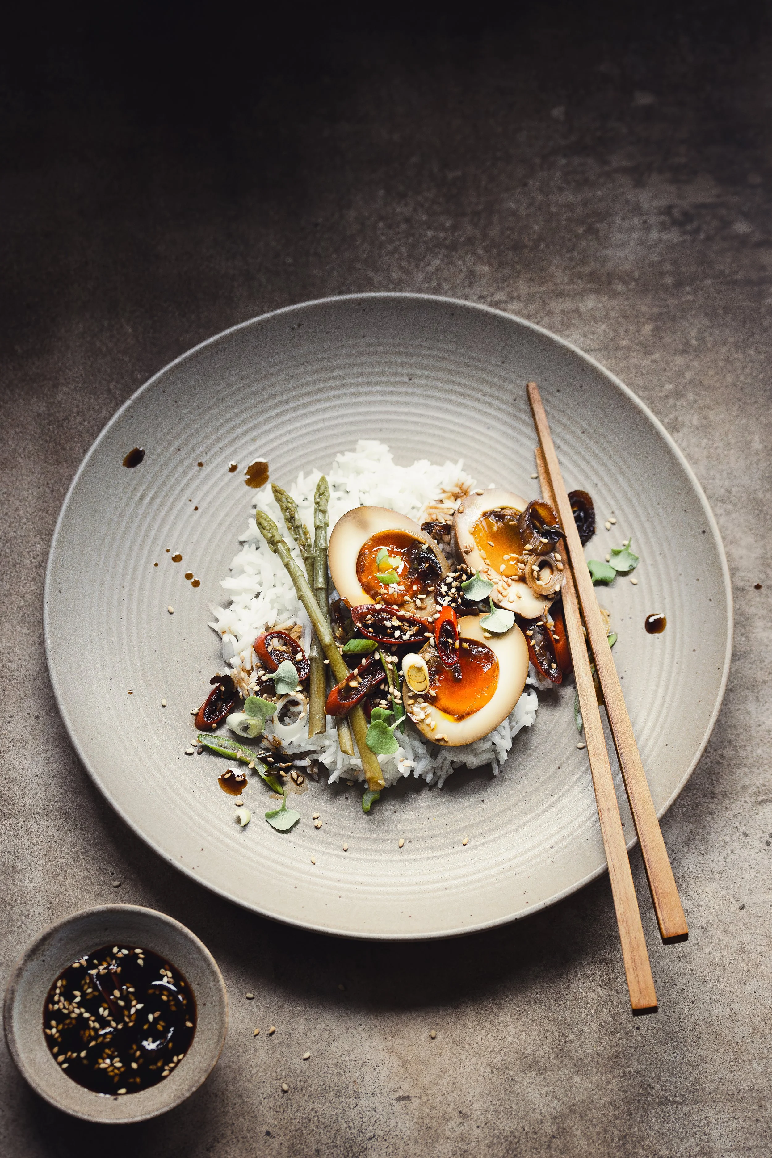 A bowl of Asian-style rice topped with halved soft-boiled eggs, sliced mushrooms, green onions, microgreens, and a soy-based sauce, with a small dish of soy sauce nearby.