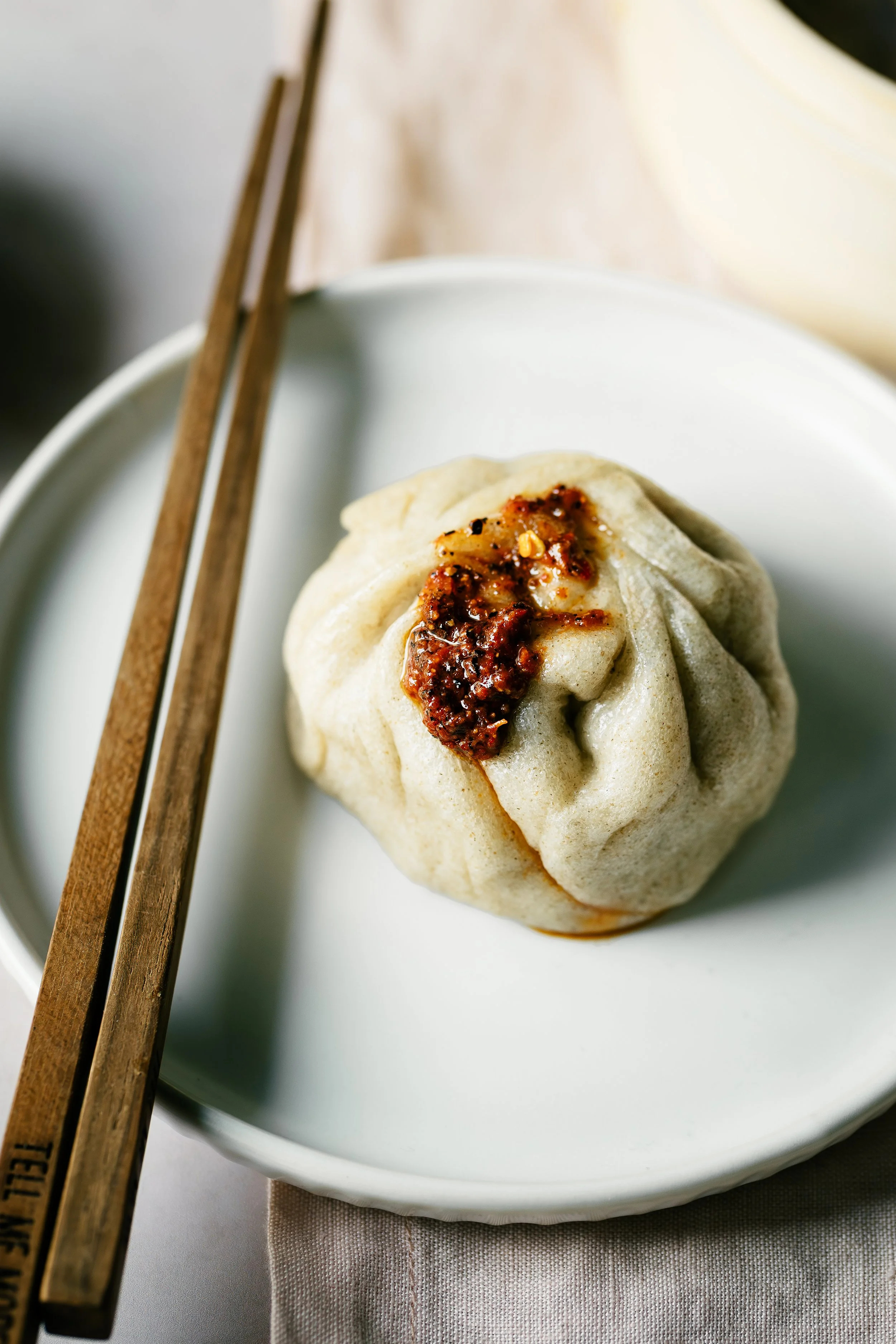 A steamed dumpling with chili oil and chili flakes on top, placed on a white plate with a spoon and chopsticks beside it.