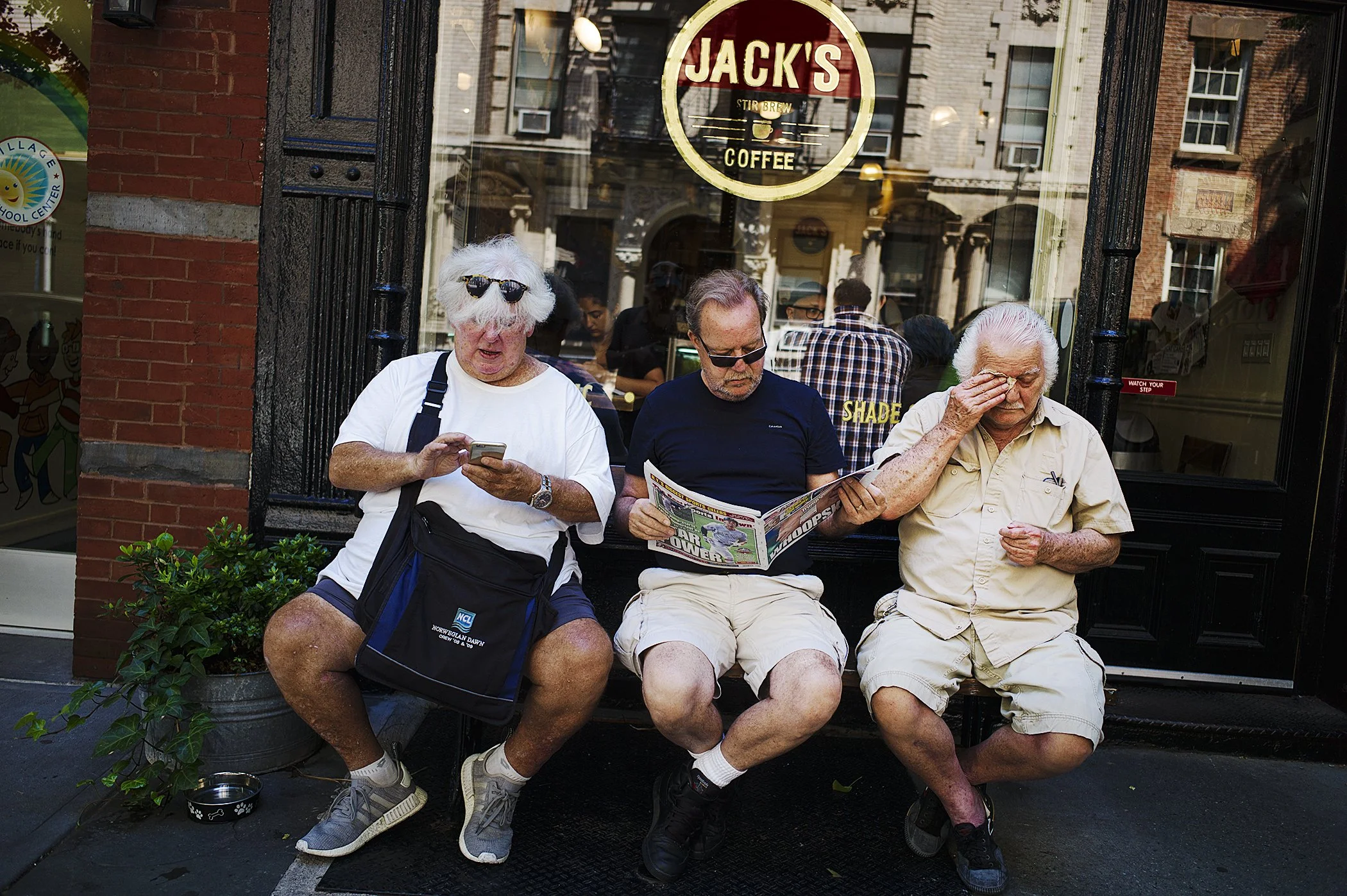 Three senior men sitting on a bench outside a coffee shop. The man on the left is looking at his phone, the middle man is reading a newspaper, and the man on the right is rubbing his eye. There is a small plant and a pet bowl near the bench. The cafe window behind them has a sign that reads 'Jack's Coffee' and shows reflections of the street.
