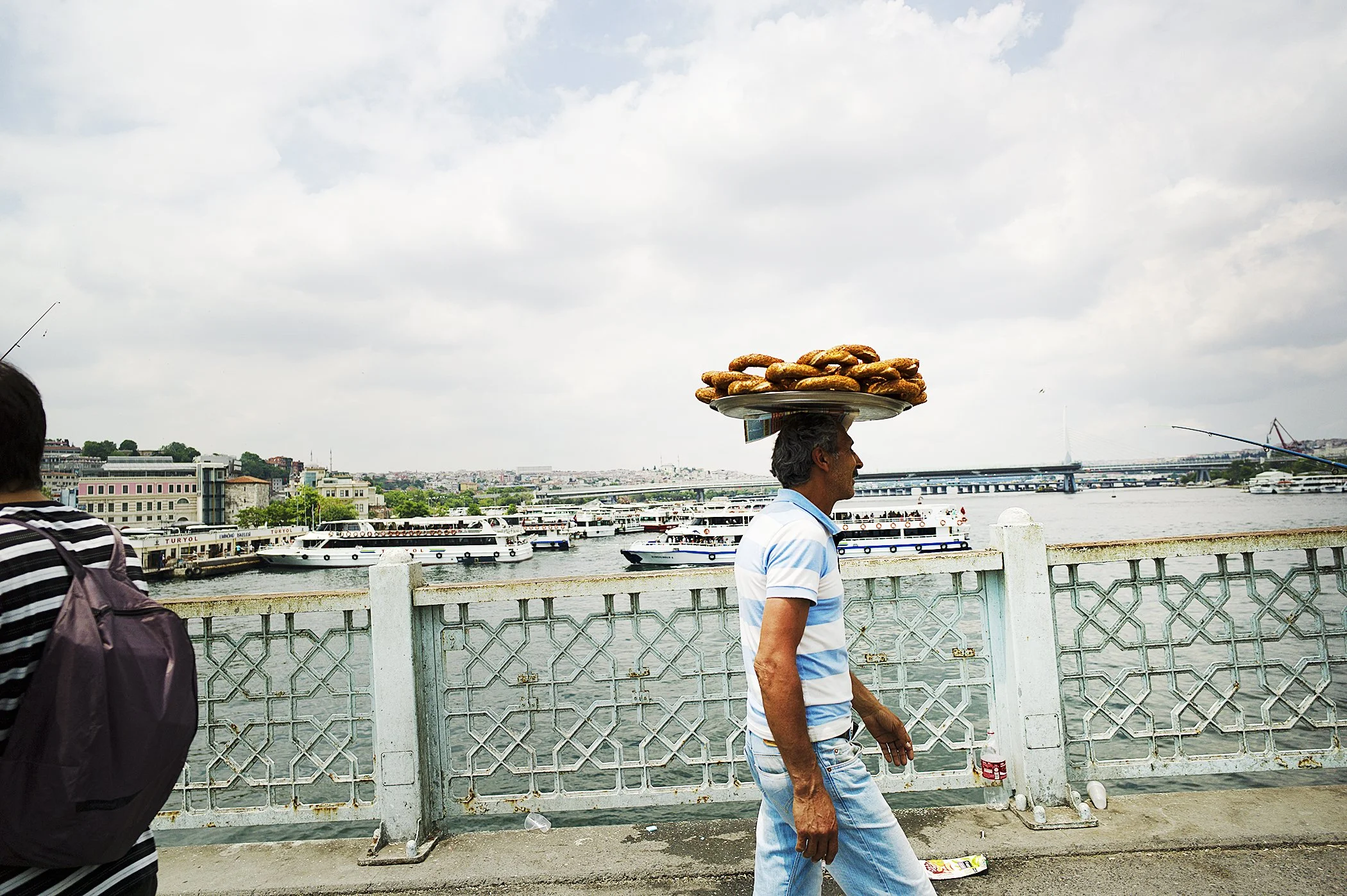 A man walking along a river promenade with his head balanced with a tray of bagels. The background shows boats docked by the river and a cloudy sky.