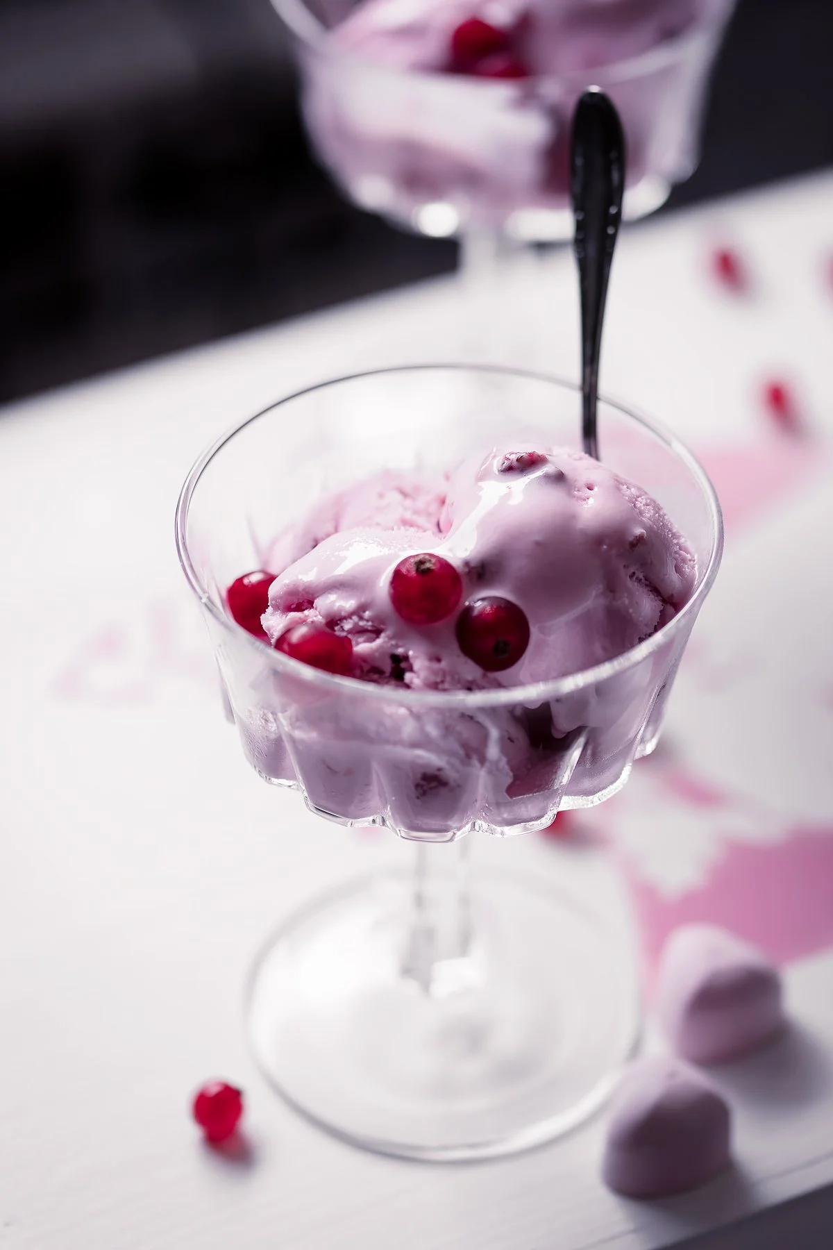 A glass dish with pink ice cream topped with red currants, on a white surface with scattered berries.