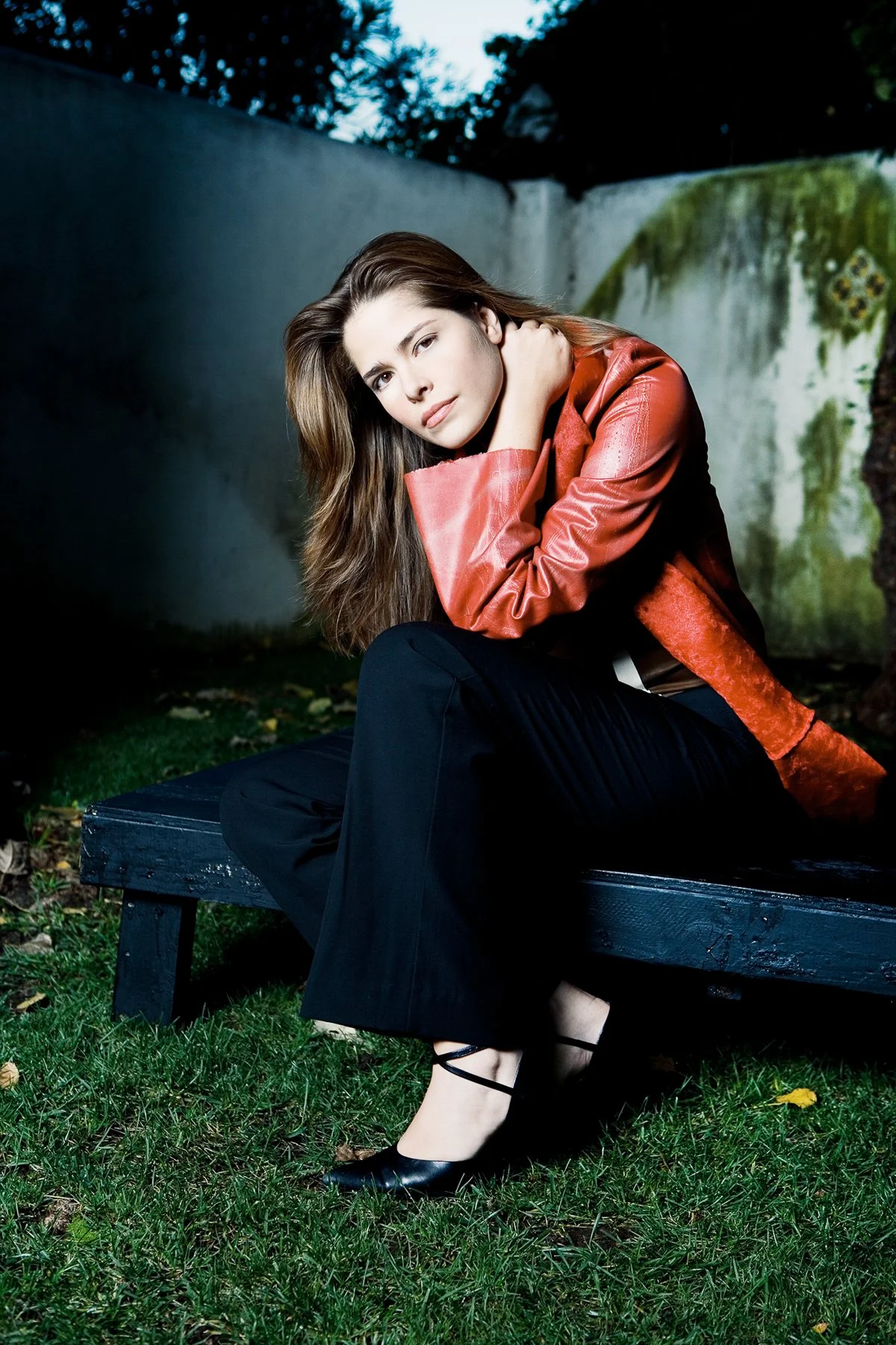 A young woman with long brown hair, wearing a red leather jacket and black wide-leg pants, sitting on a black bench outdoors next to a mossy wall, looking at the camera.