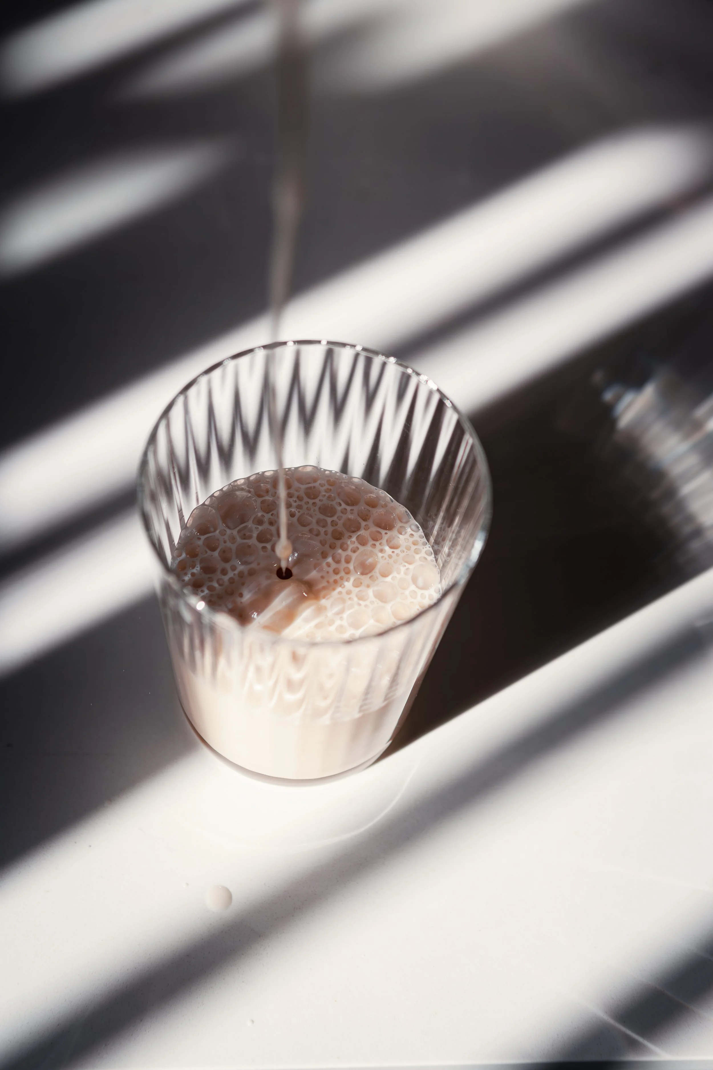 A glass with bubbly of a light-colored beverage being poured, with shadows of window blinds cast across the scene.