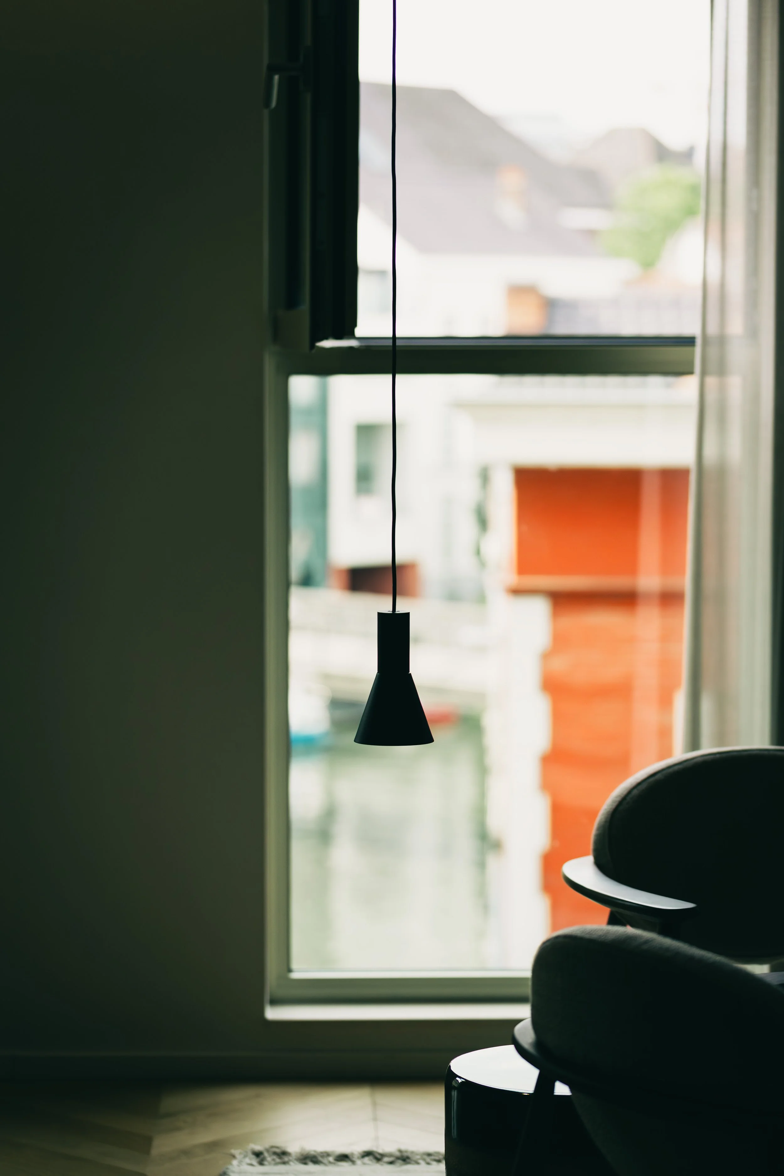 A view of a modern interior with a large window, a black hanging pendant light, and a dark-colored chair or sofa near the window.